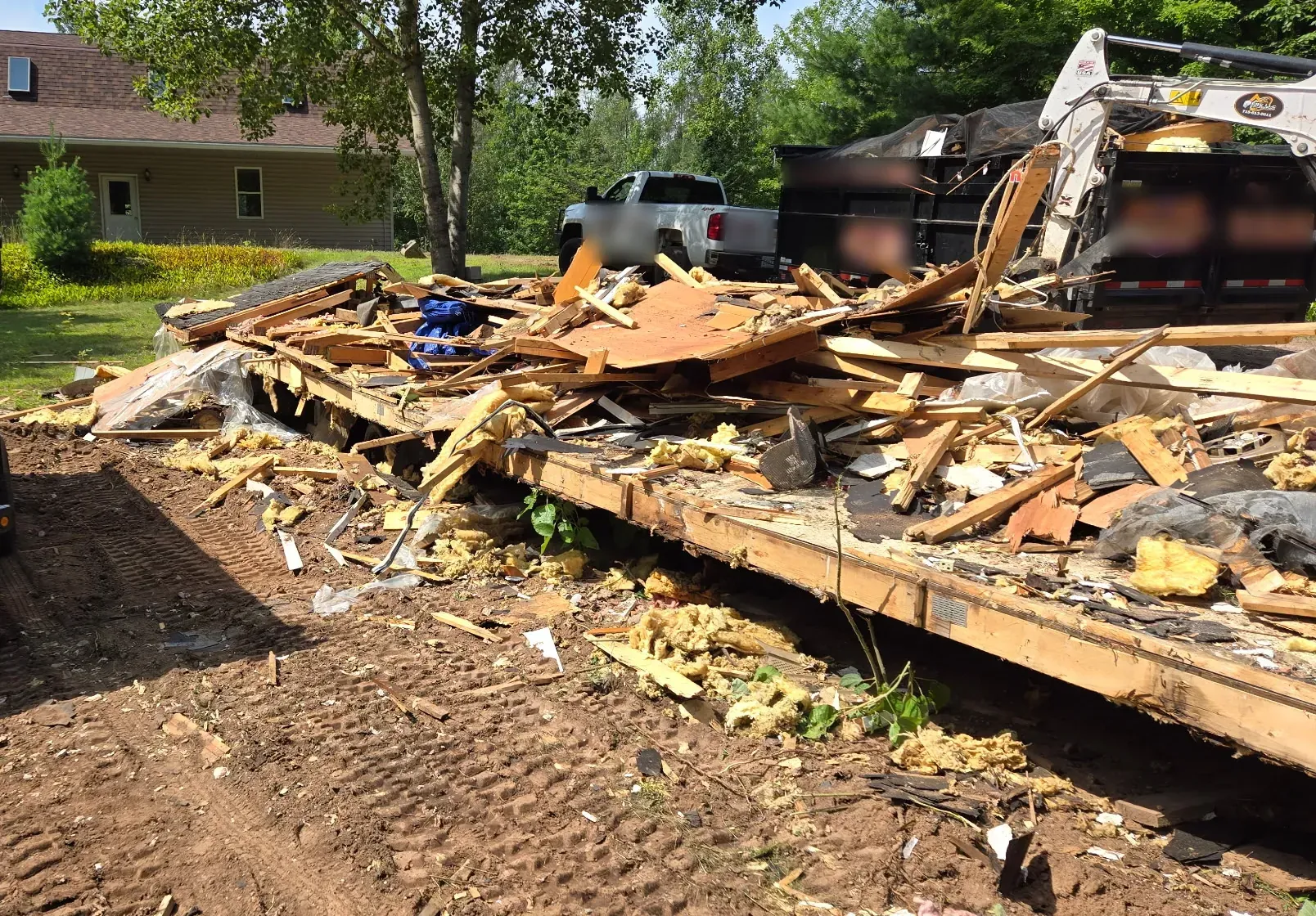Mini excavator tearing down a mobile home, with the collapsed floor deck and framing debris loaded toward roll-off dumpsters in Phillips, WI