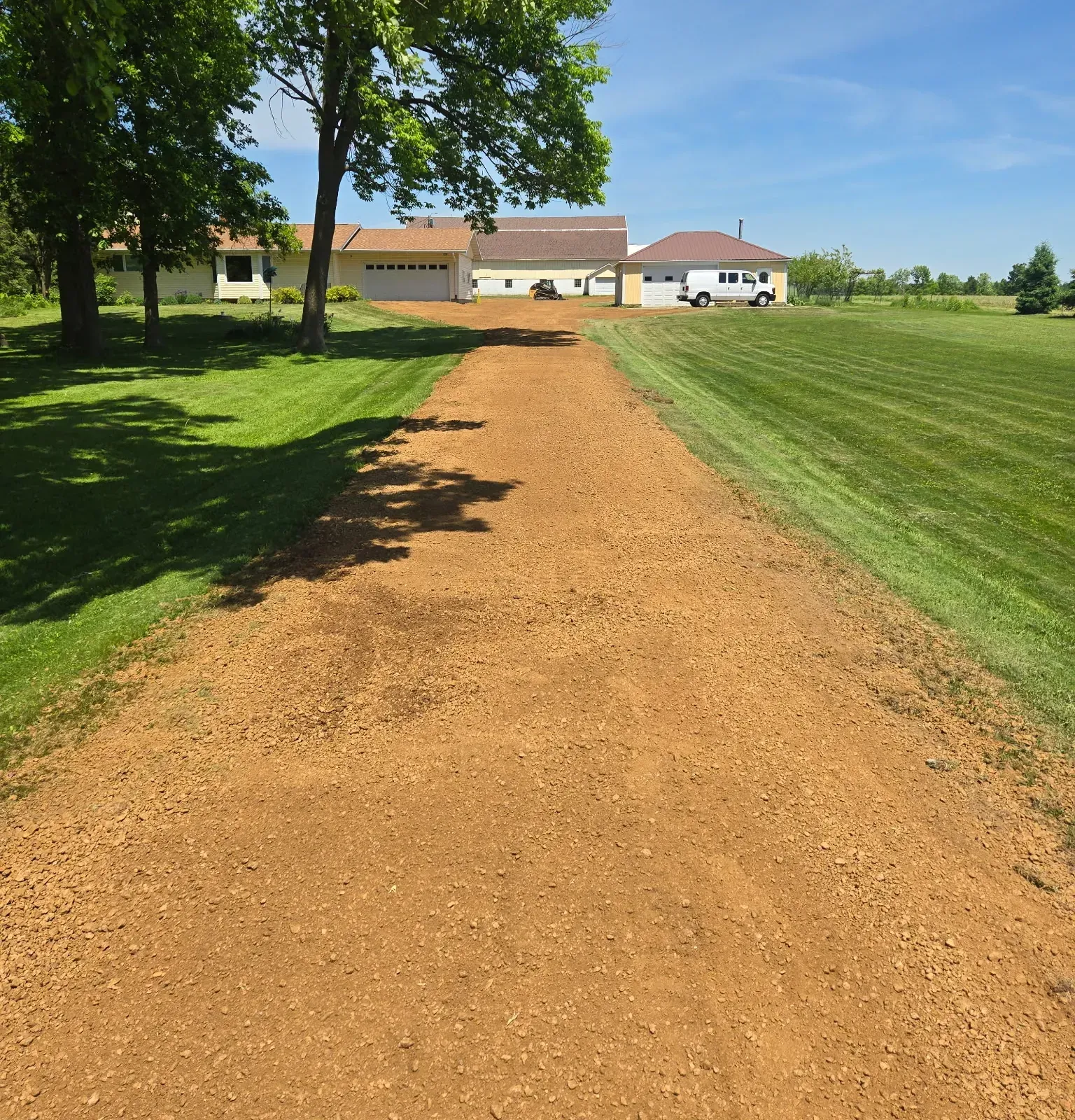 Long farm driveway top dressed with new red crushed gravel between mowed lawns leading to the house and garages in Spencer, WI
