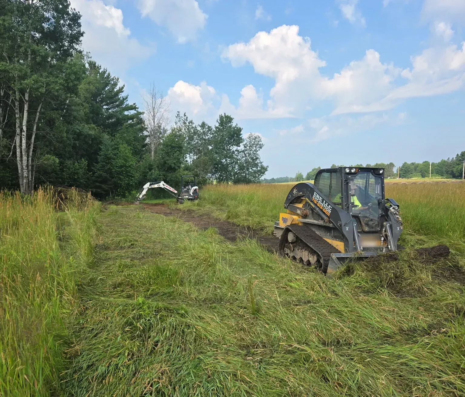 ERE track loader and mini excavator digging a drainage ditch through tall grass along a wooded field edge in Rib Lake, WI