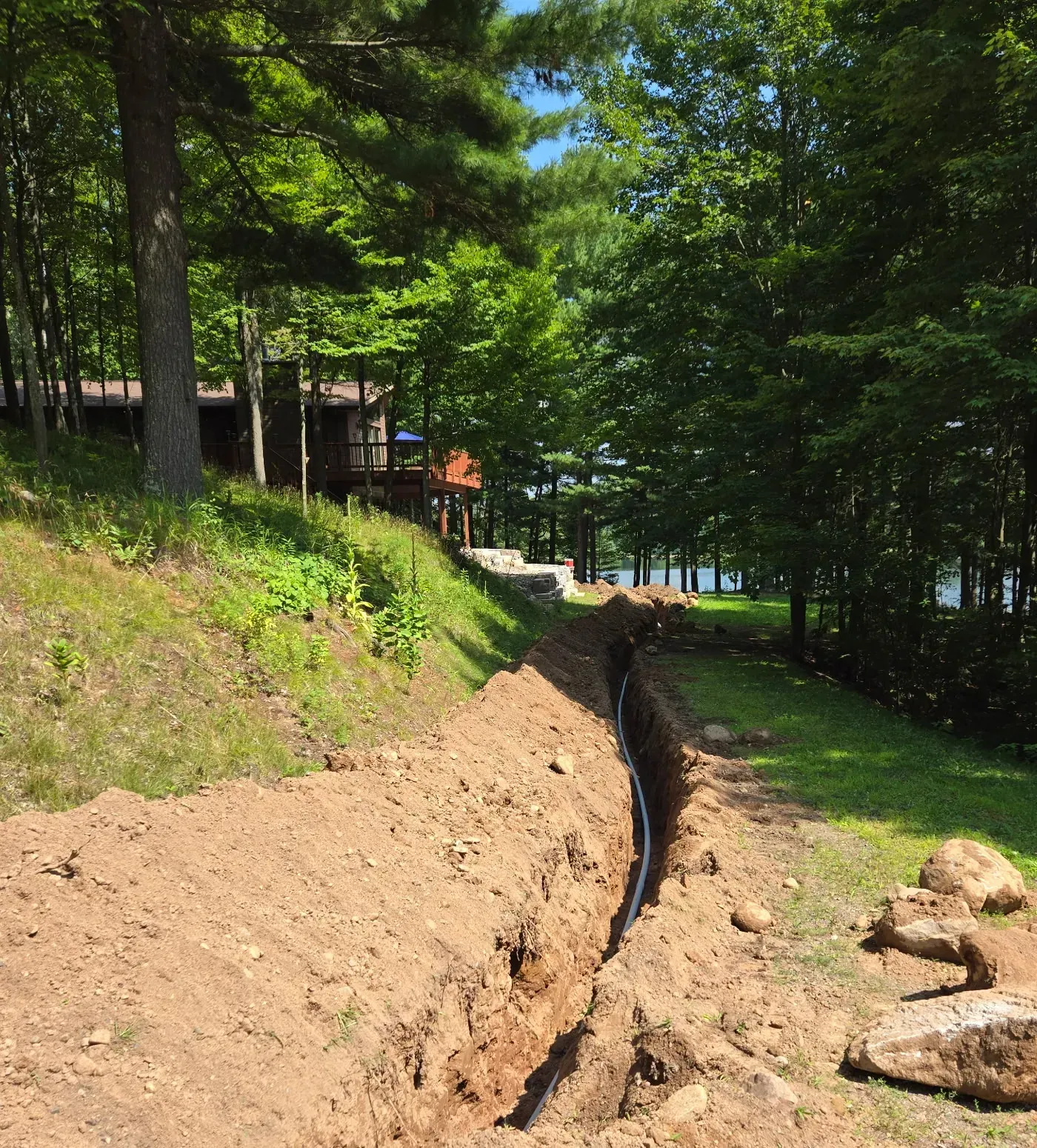 Long excavated trench carrying underground power conduit past a hillside cabin toward the lake in Phillips, WI