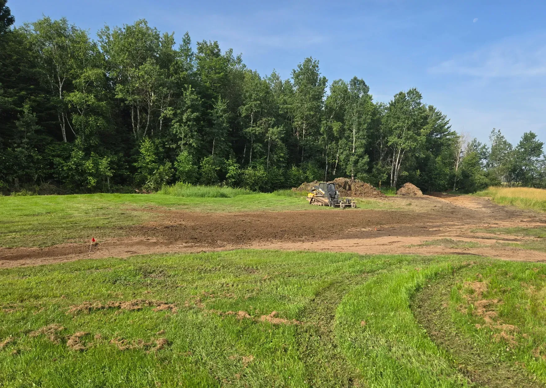 Regraded yard with a shaped drainage swale marked by flags, and a track loader with a grading rake beside spoil piles in Rib Lake, WI