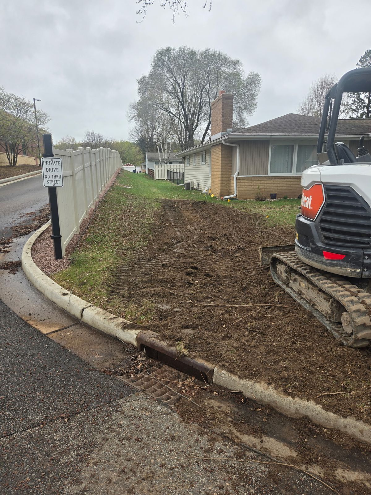 Mini excavator regrading a side yard so water drains away from the house foundation and toward the street storm drain in Wausau, WI