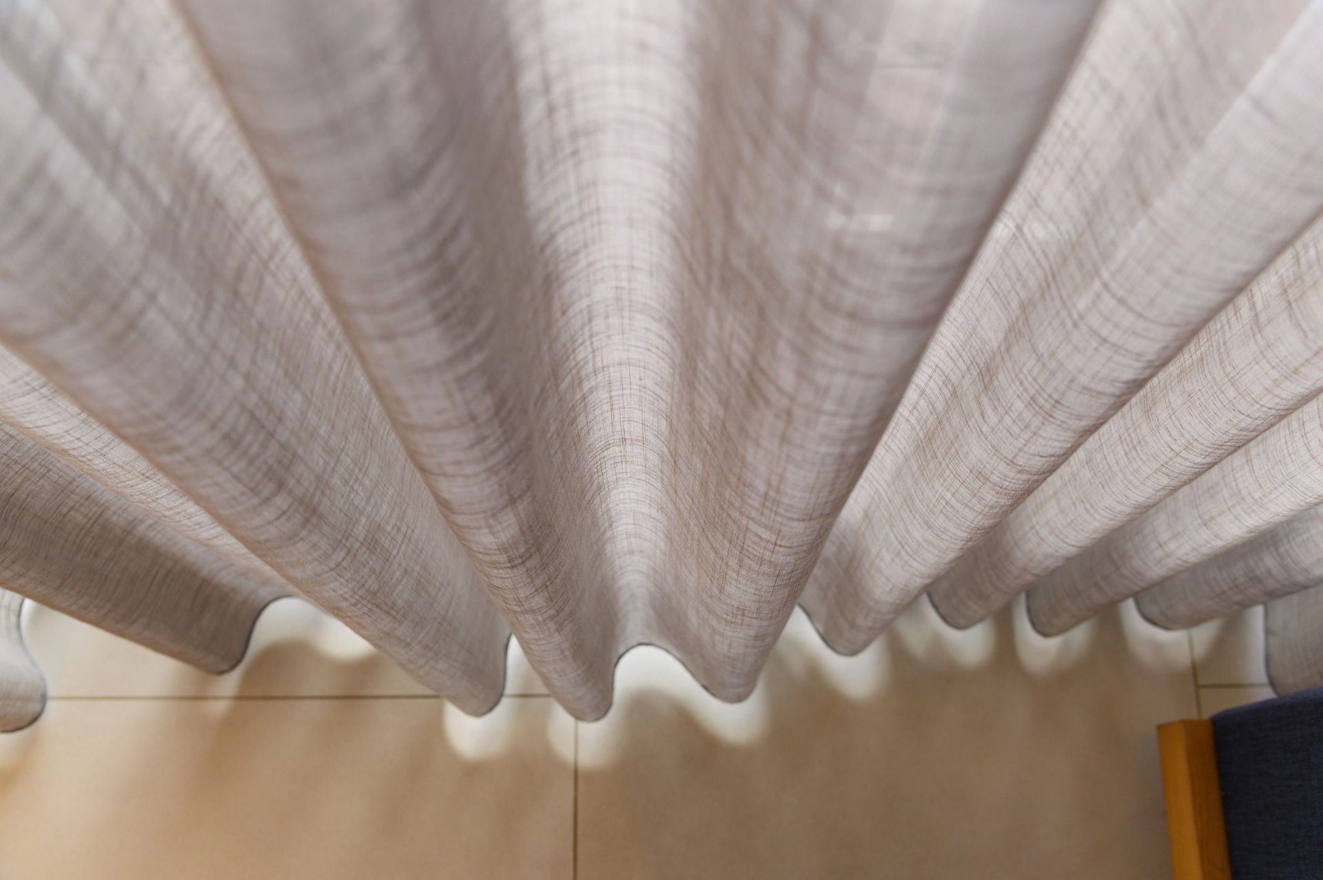 Tende beige che scendono su un pavimento piastrellato, con uno scorcio di mobili blu sul lato destro.