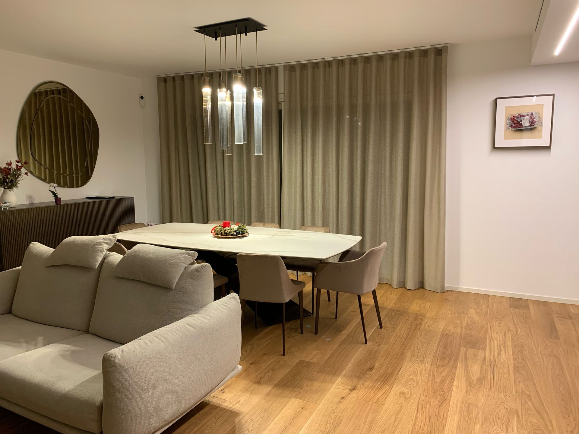 Sala da pranzo e soggiorno moderni con divano beige, pavimento in legno, tavolo rotondo e tende.