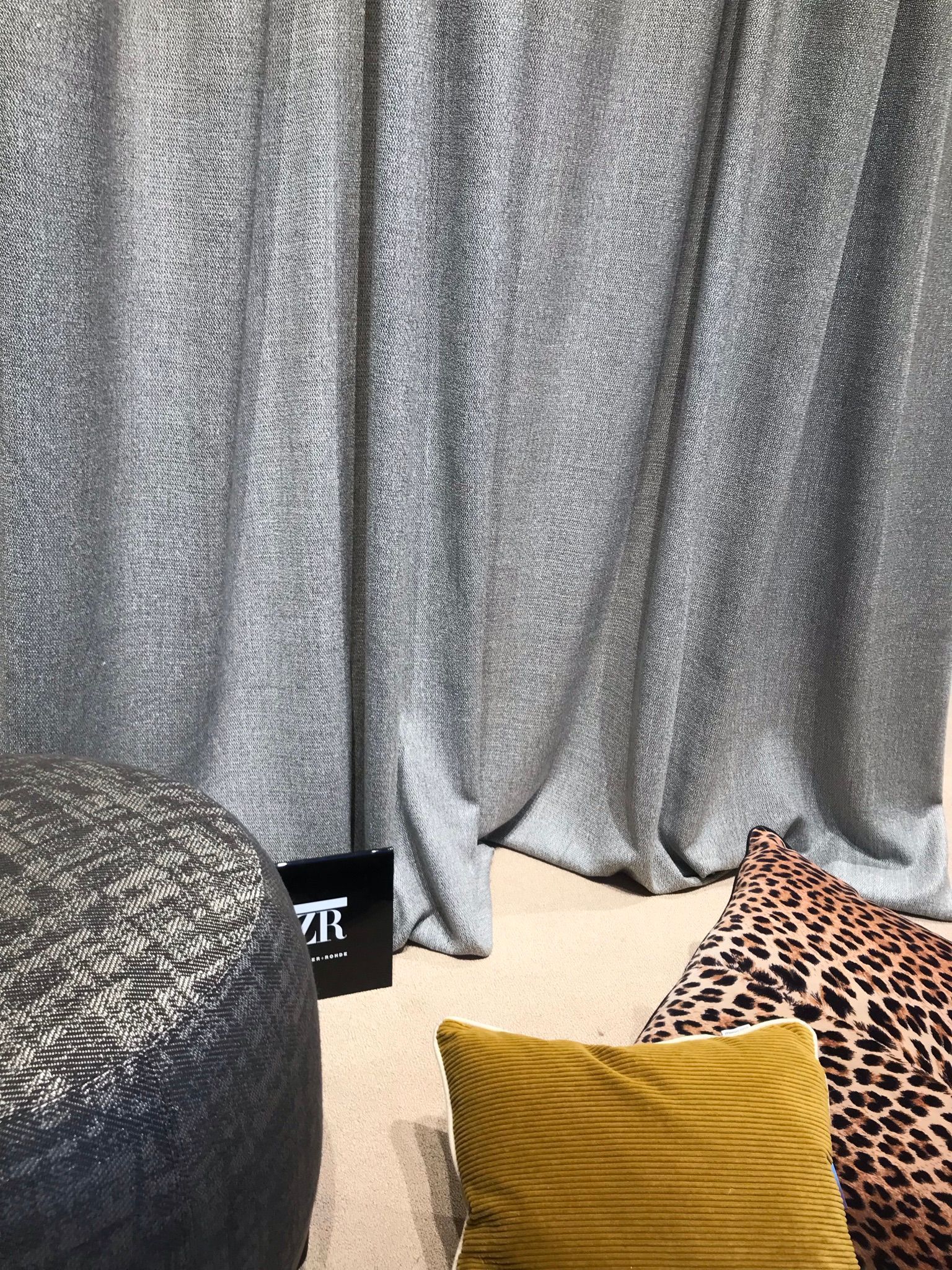 Tende argentate dietro un divano beige con cuscini leopardati e color senape, accanto a un pouf nero.