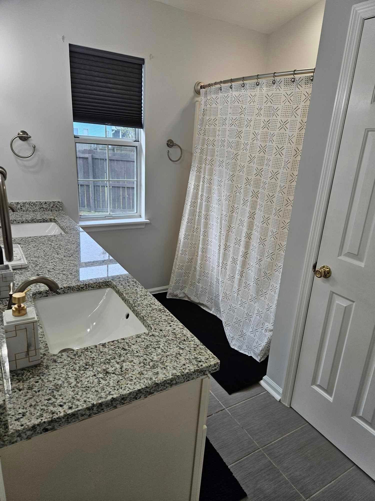 Bathroom with double sinks, granite countertops, shower with curtain, window with blind.