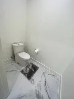 Toilet in a small bathroom with marble-look flooring, toilet paper holder, and a small rug.