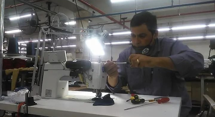 Un técnico en un taller de fábrica repara una máquina de coser industrial con un destornillador bajo una lámpara brillante.