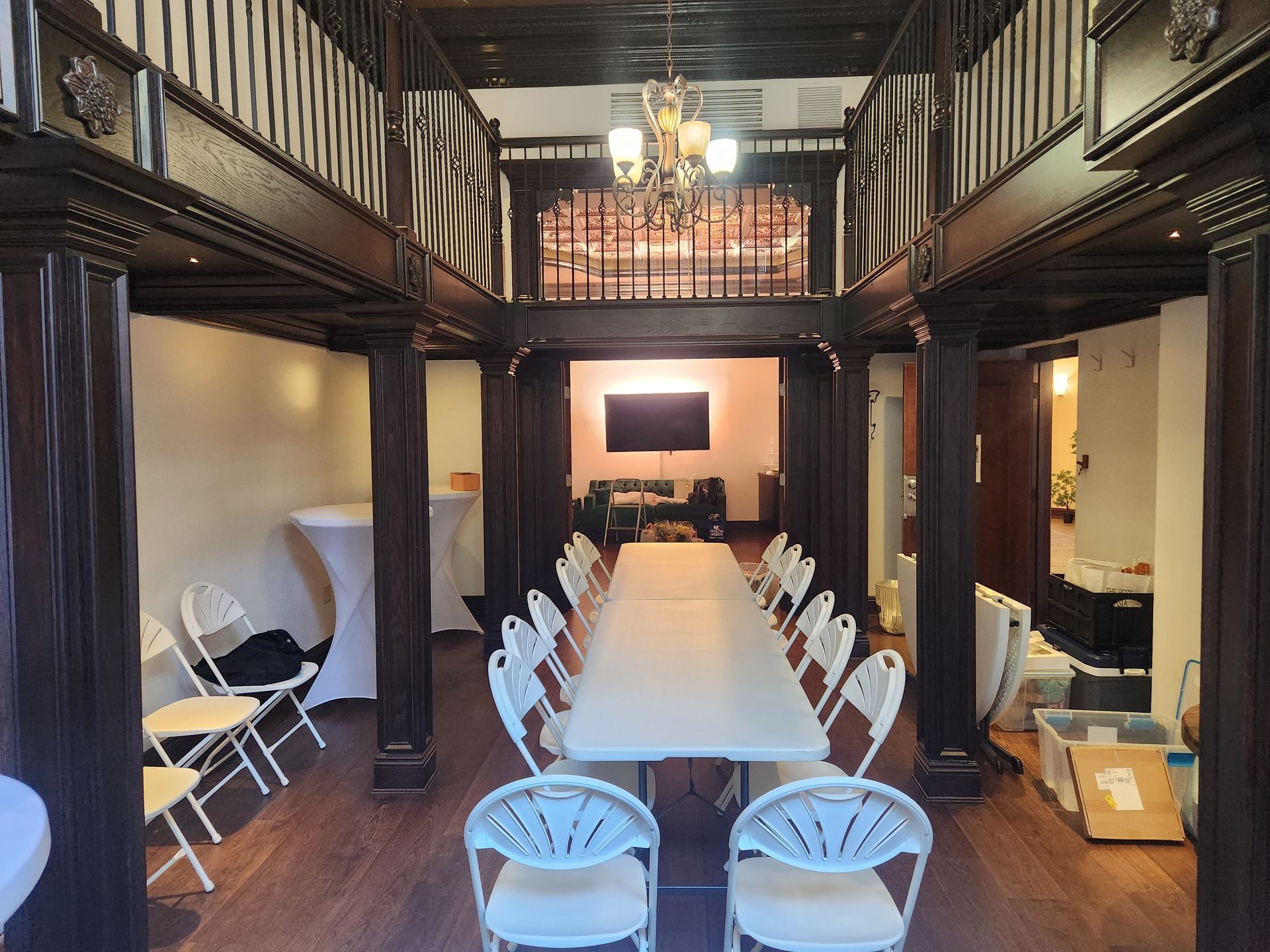 Long rectangular table set for a gathering in a dark-wood paneled room with white chairs. A TV hangs in the background.
