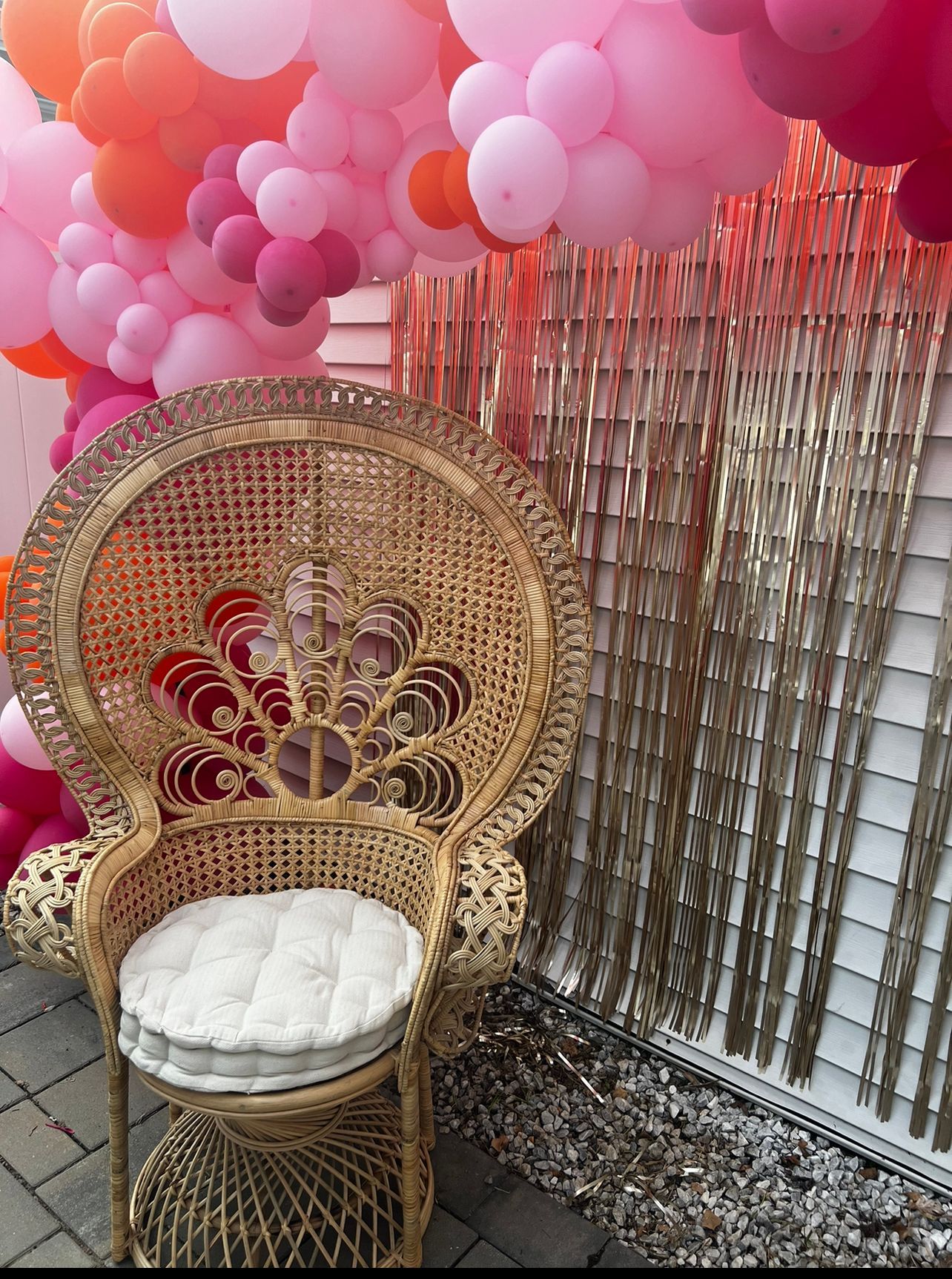 Wicker peacock chair with white cushion under a pink and orange balloon arch, next to gold fringe wall.