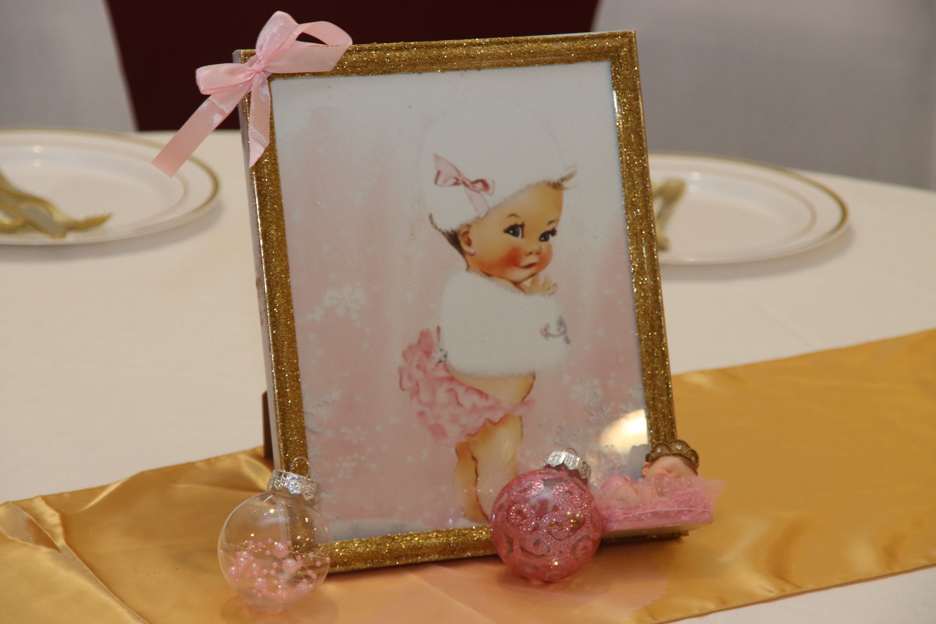 Baby shower centerpiece: Framed vintage illustration of a baby in pink tutu; gold-trimmed frame, pink ribbon, glitter ornaments, gold table runner.
