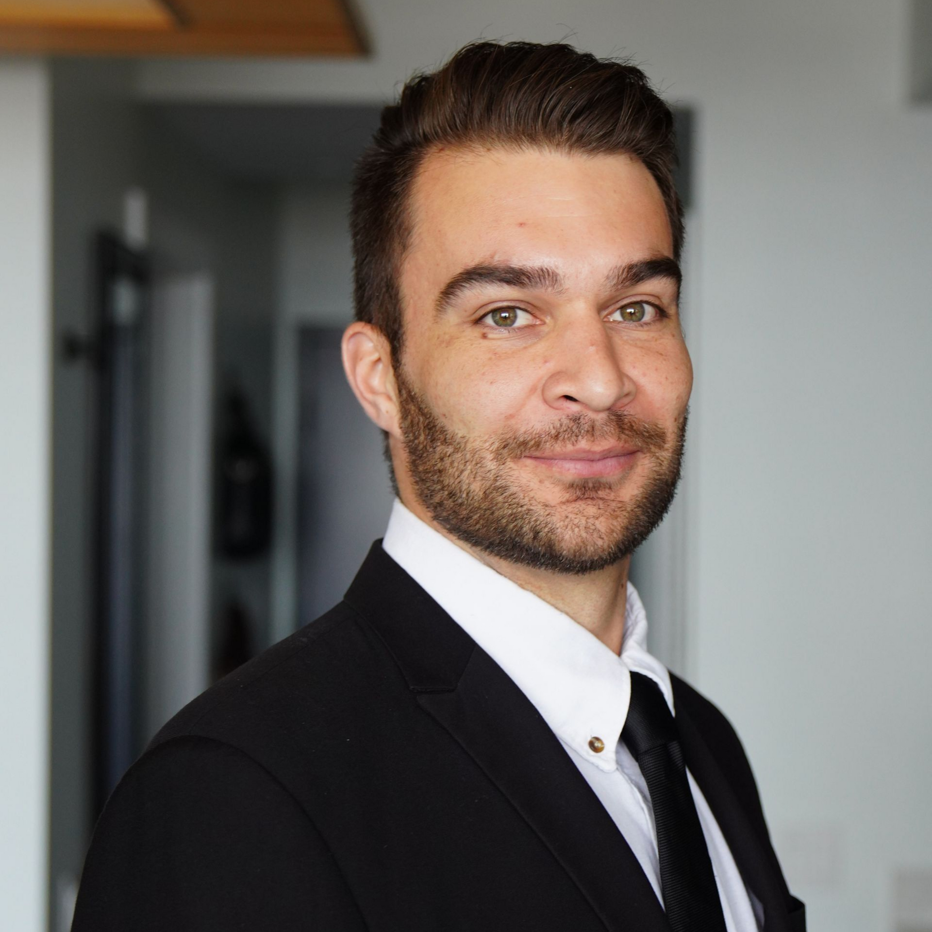 Man in black suit and tie, smiling, looking at the camera. Indoor setting, light background.