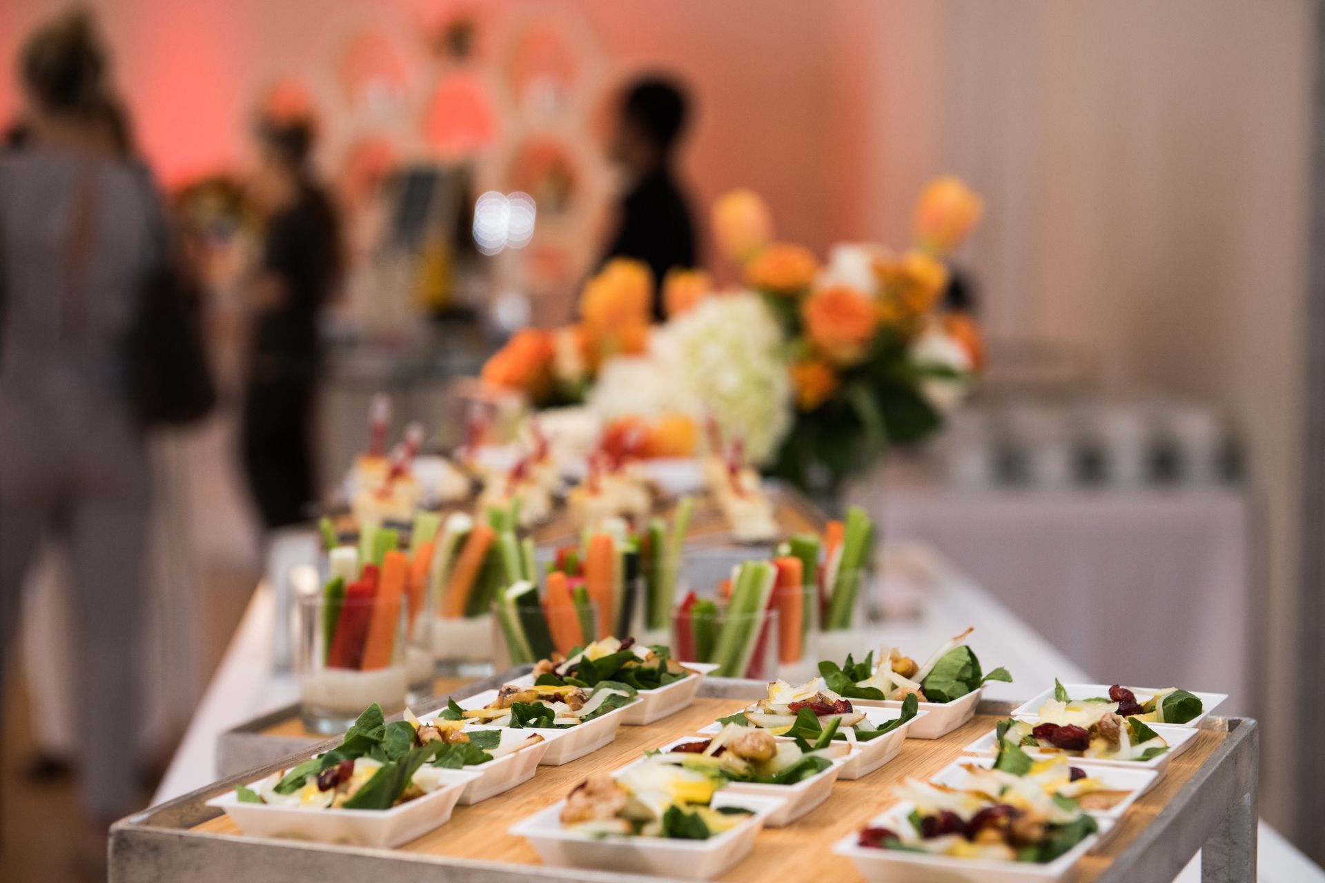 Appetizers on a buffet table: vegetables, salads, and flowers; people blurred in the background.