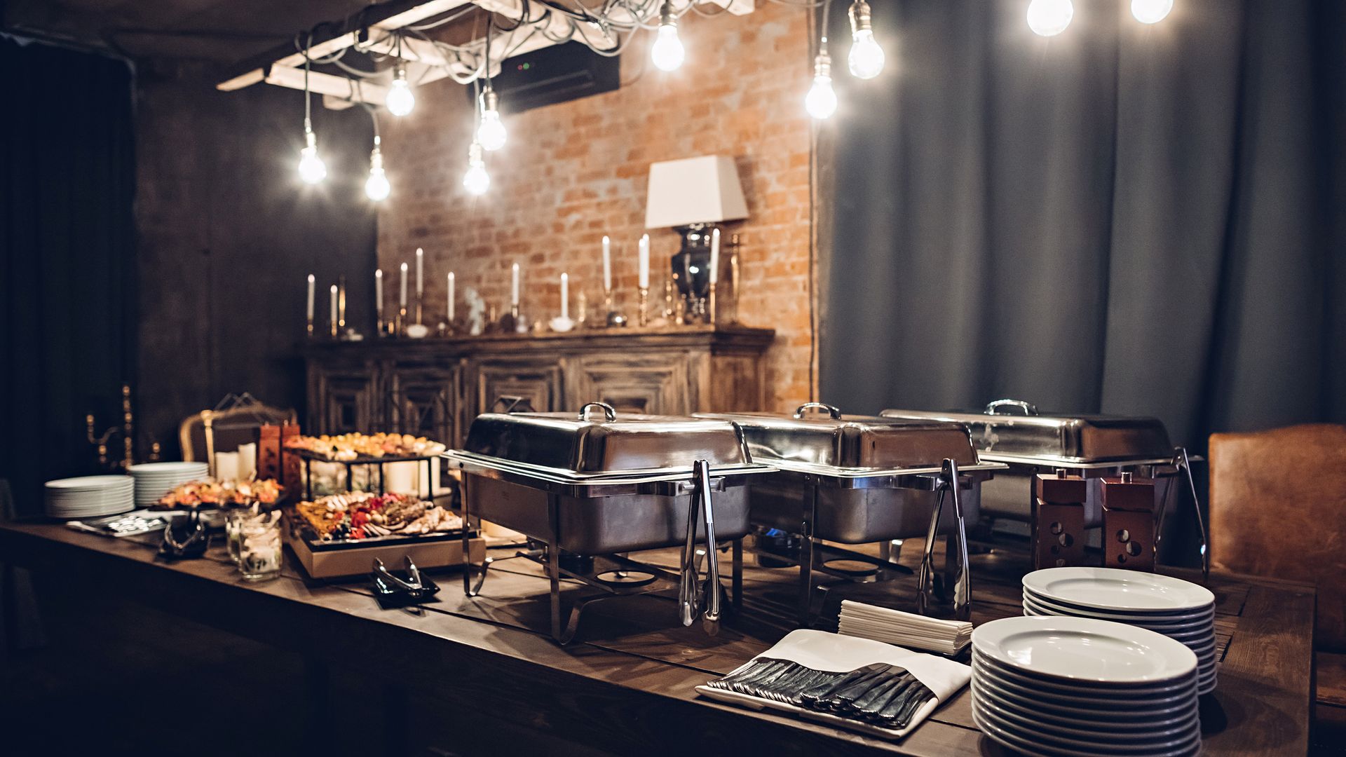 Buffet table with food and serving dishes in a dimly lit room with exposed brick and decorative lighting.