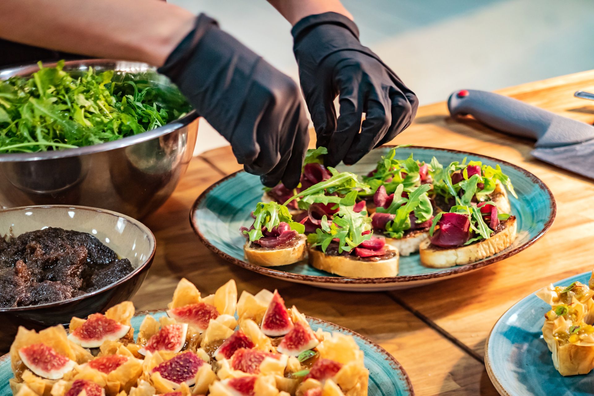 Chef in black gloves garnishes toasted bread with arugula and beetroot on a plate, table setting.
