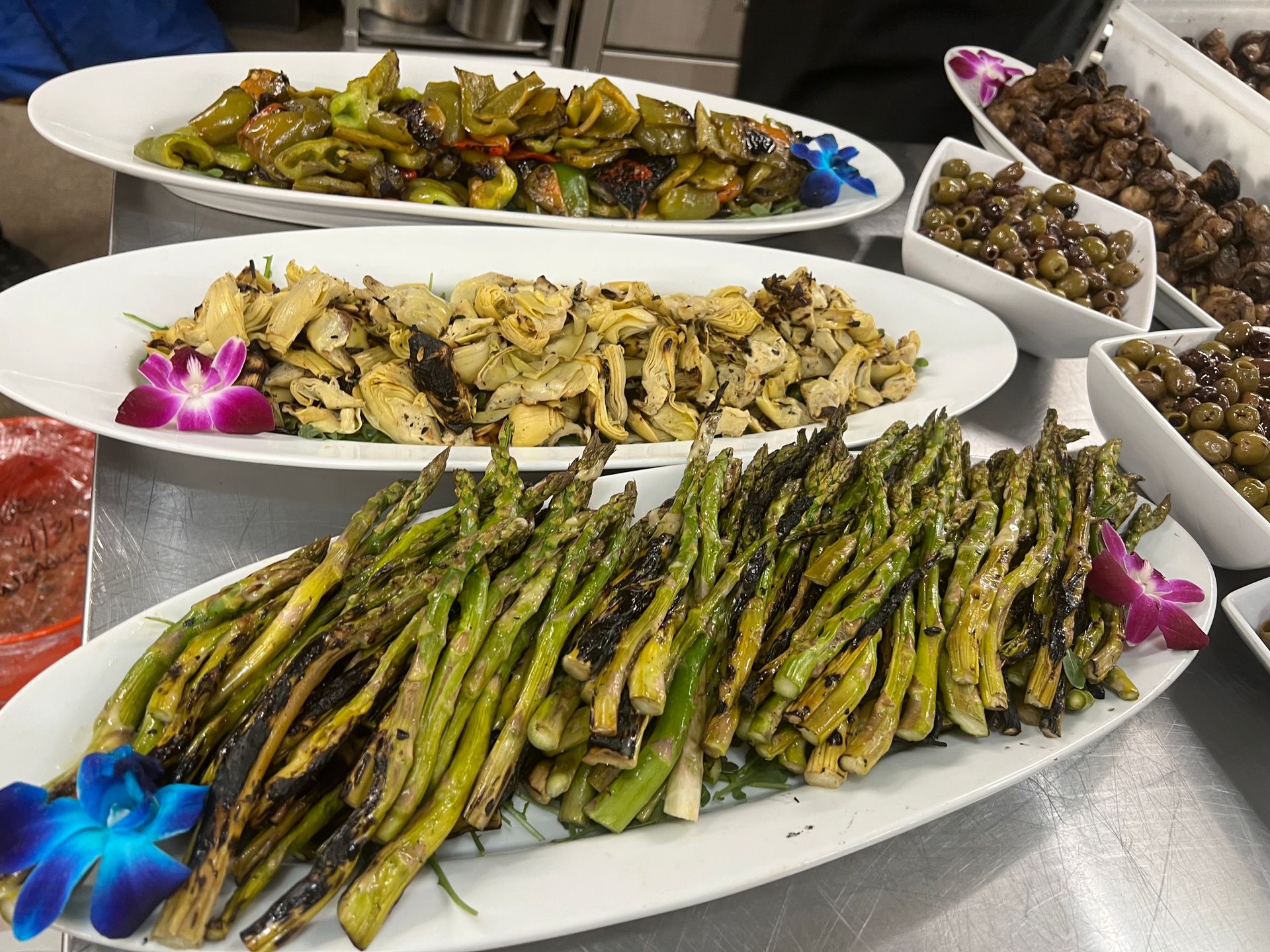 Three oblong white platters of roasted vegetables, including asparagus and artichokes, arranged on a metallic counter.