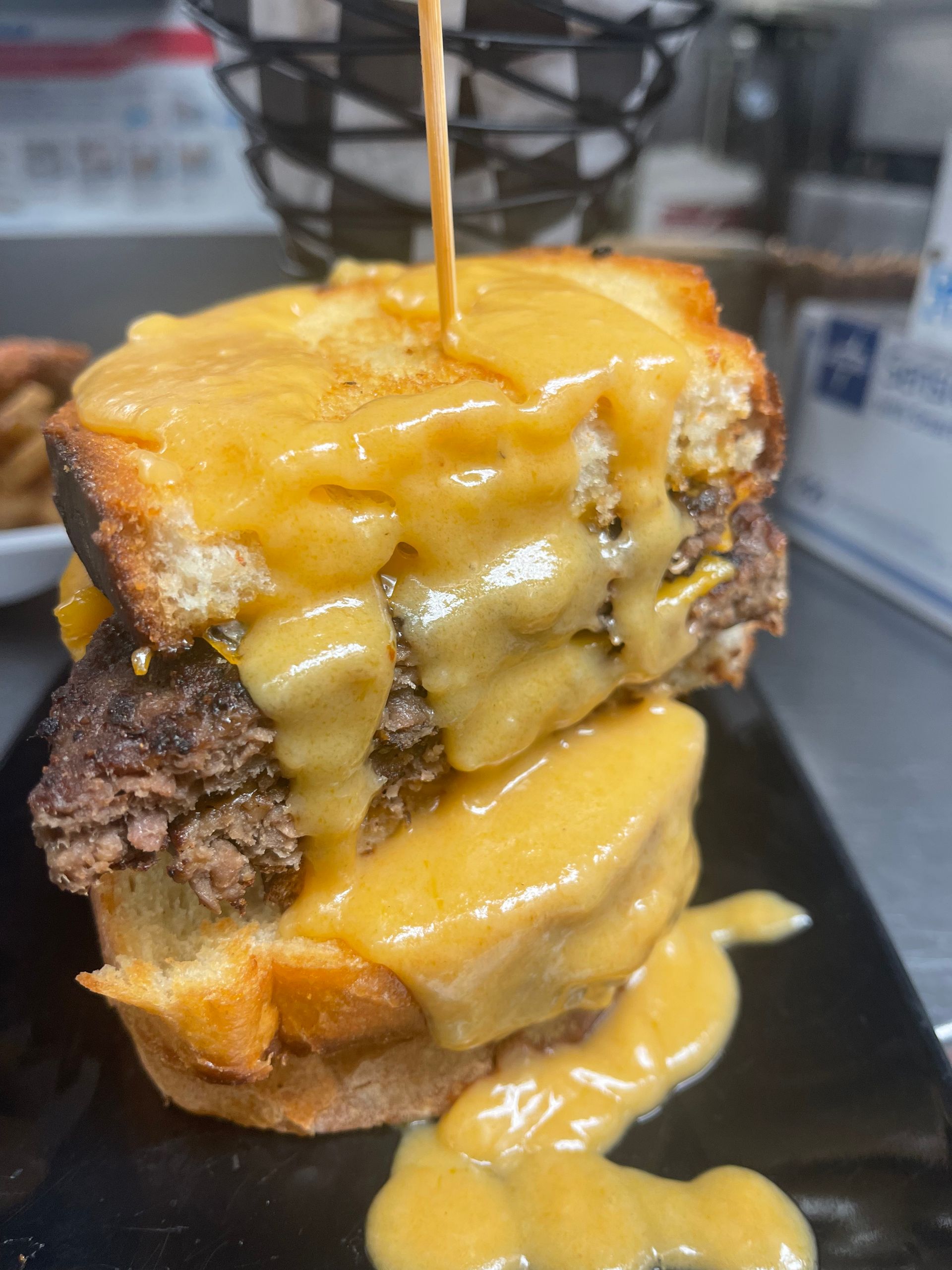 A double-decker burger on toasted bread covered in thick, melted orange cheese, with sauce dripping down onto a black plate.