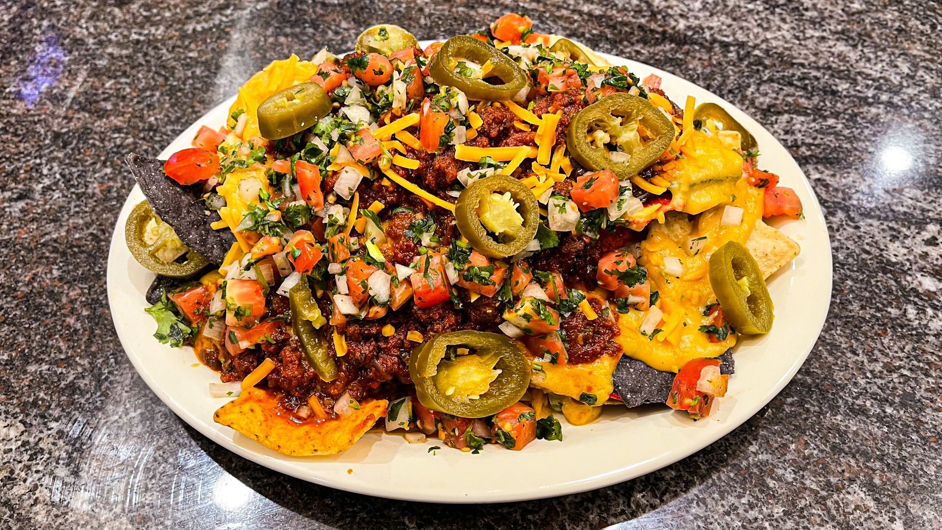 A plate of nachos topped with melted cheese, diced tomatoes, onions, cilantro, and sliced jalapeños on a stone surface.