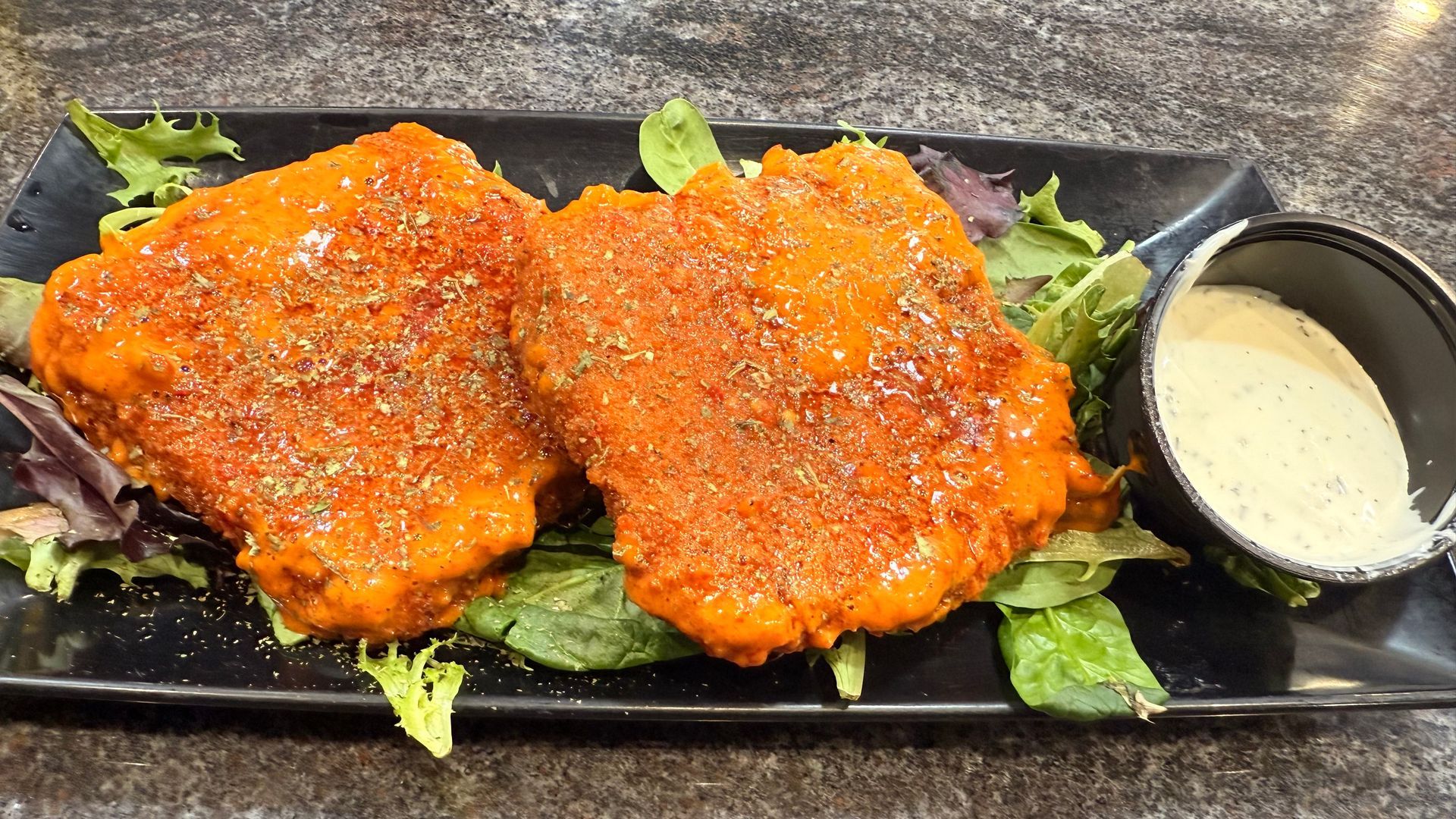 Two breaded, buffalo-sauced chicken cutlets served over a bed of greens with a side of ranch dressing on a black tray.