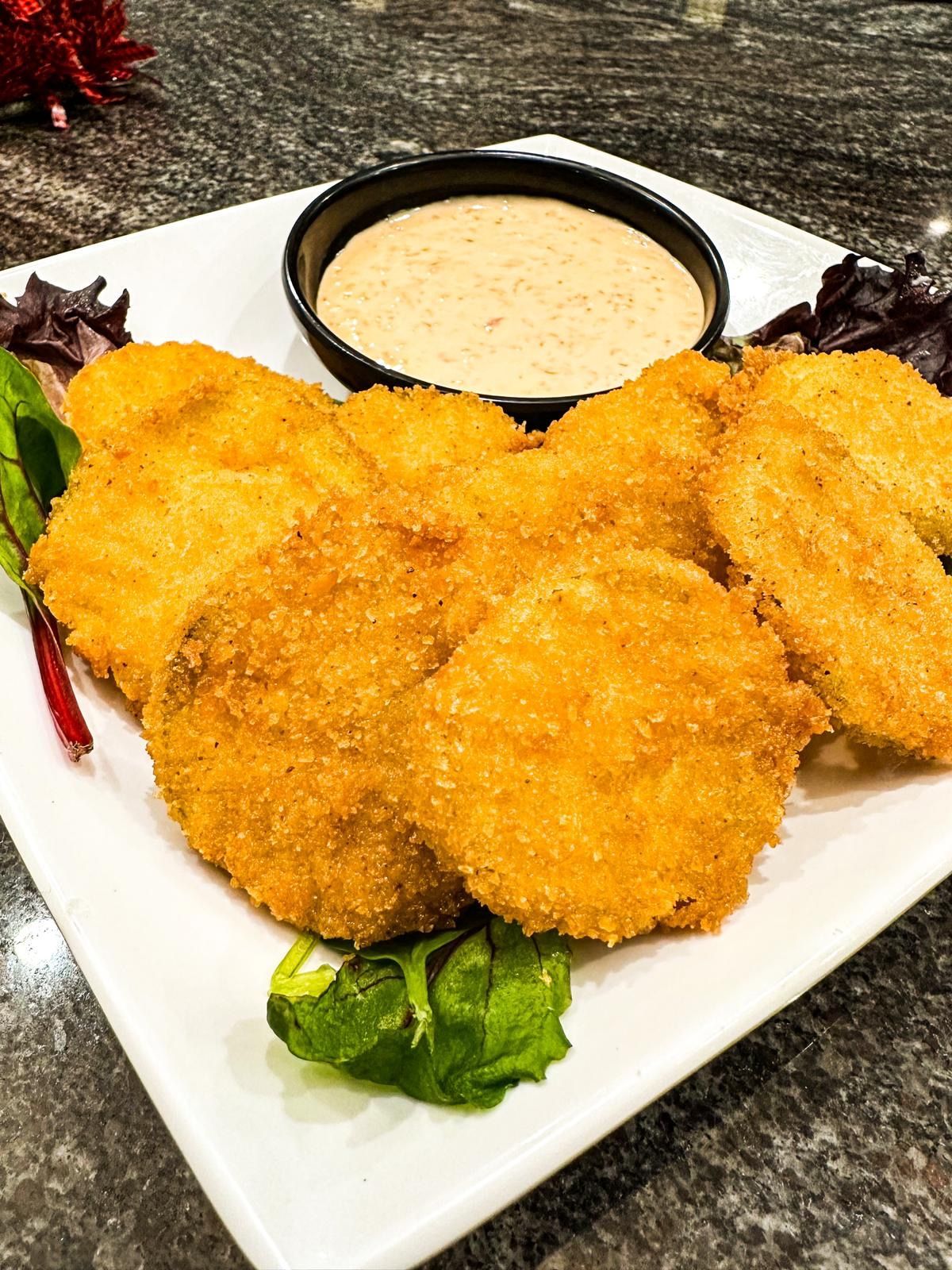 Fried green tomatoes served on a white plate with a side of dipping sauce and garnished with greens.
