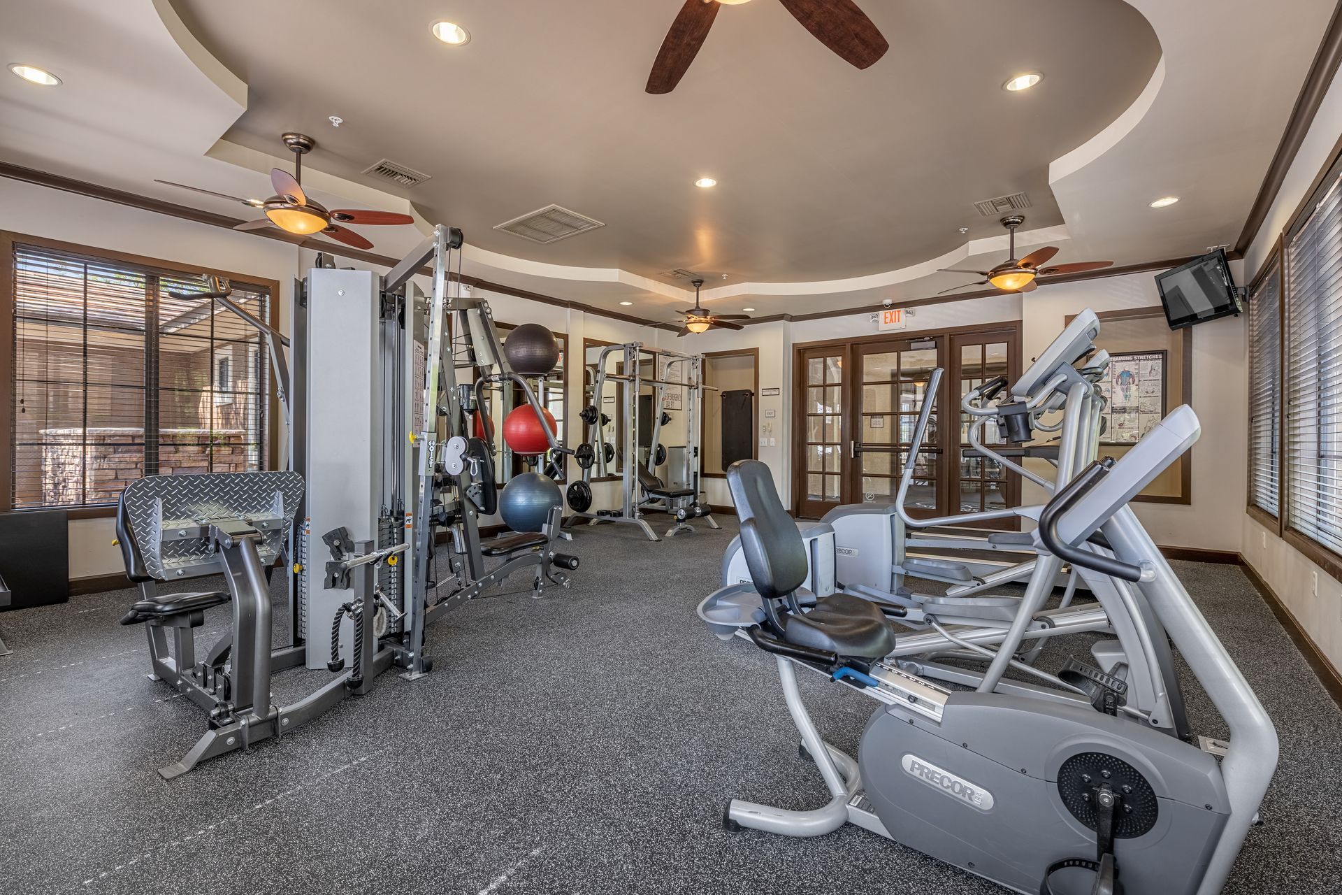 Home gym with cardio and weight machines in a bright room with large windows and ceiling fans