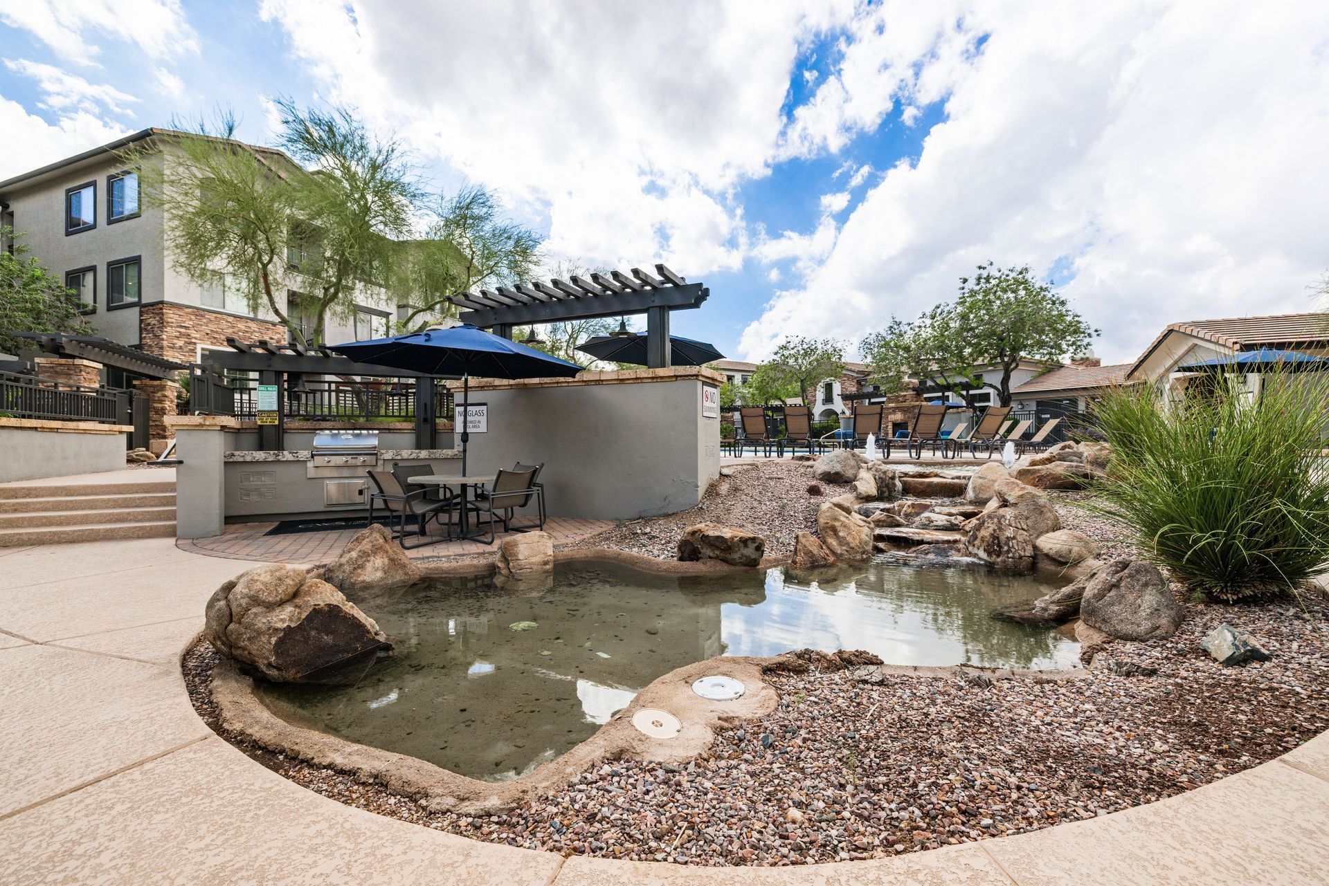 Outdoor courtyard with a small pond, rocks, patio seating, and a pergola under a partly cloudy sky