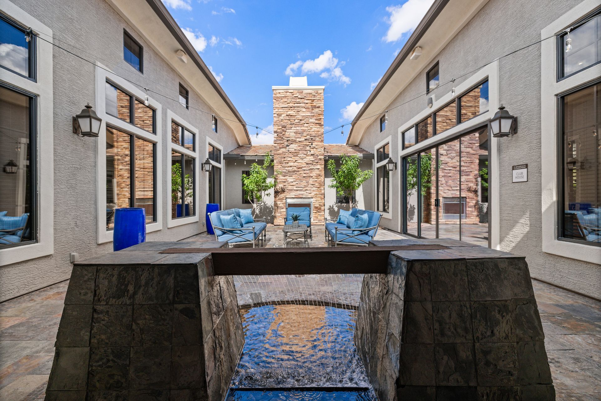 Outdoor patio between two buildings with a stone fireplace and blue-cushioned chairs around a table