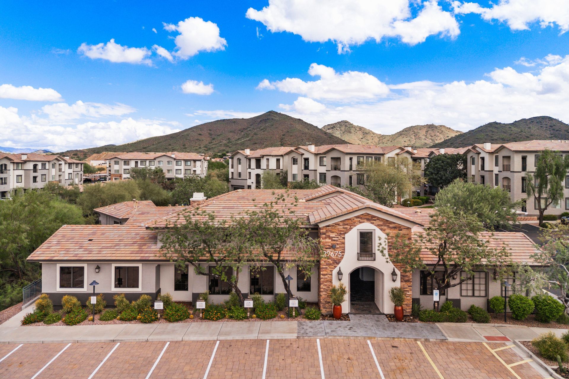 Aerial view of a Mediterranean-style apartment complex with red roofs, mountains, and a blue sky.