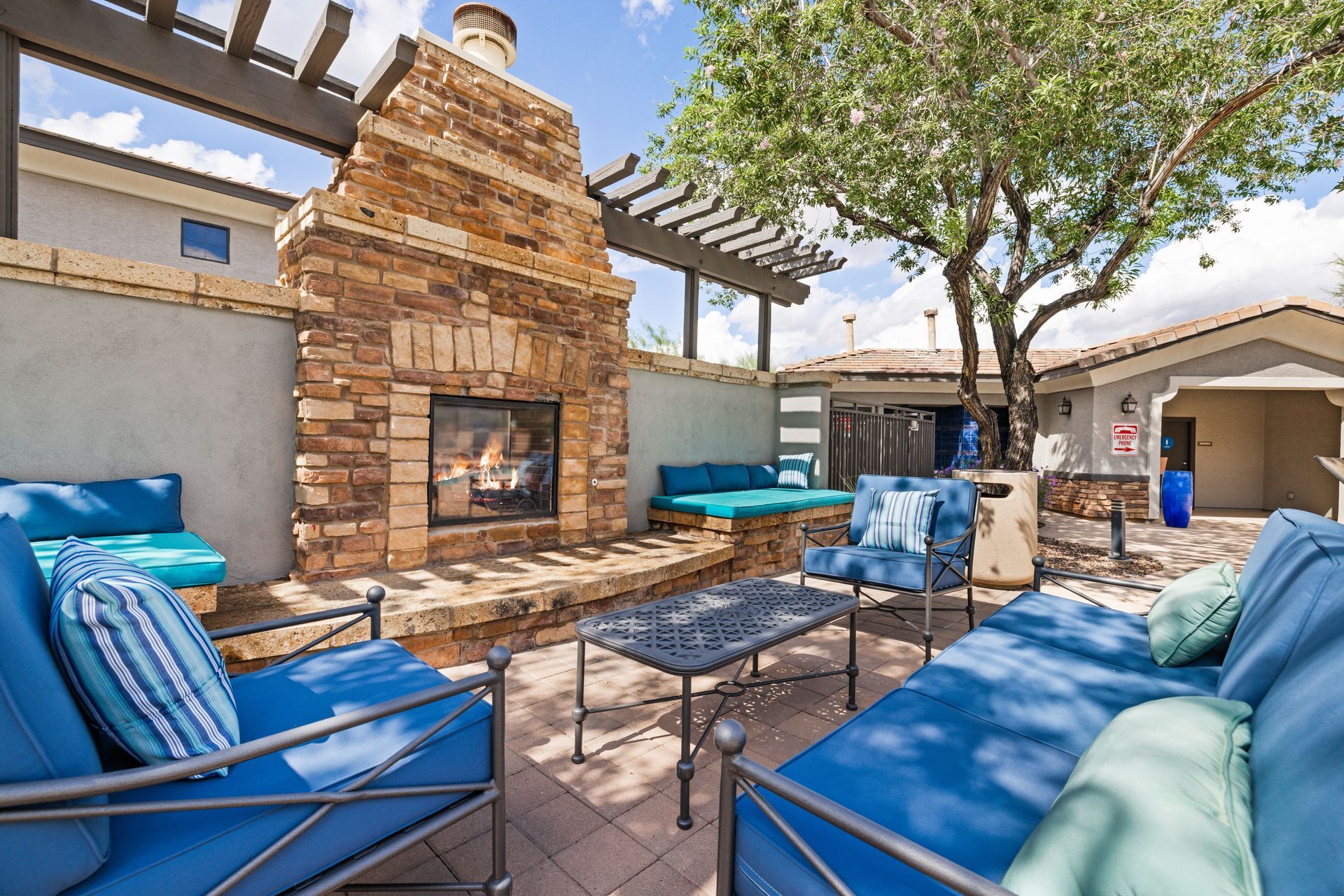 Patio with blue lounge chairs and cushions around a stone fireplace under a pergola, beside a tree.