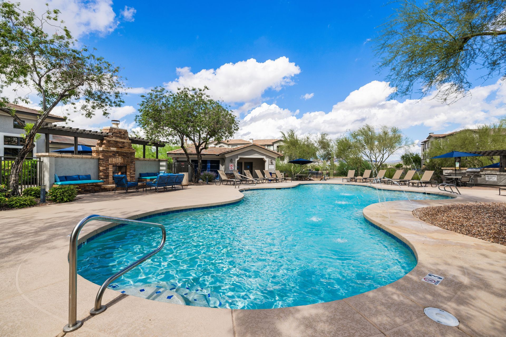 Outdoor resort pool with turquoise water, lounge chairs, trees, and blue sky with clouds