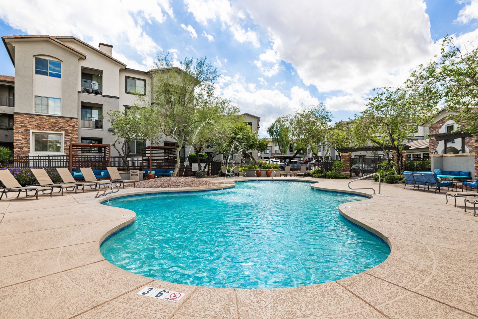 Apartment courtyard pool with blue water, lounge chairs, and surrounding buildings under a partly cloudy sky