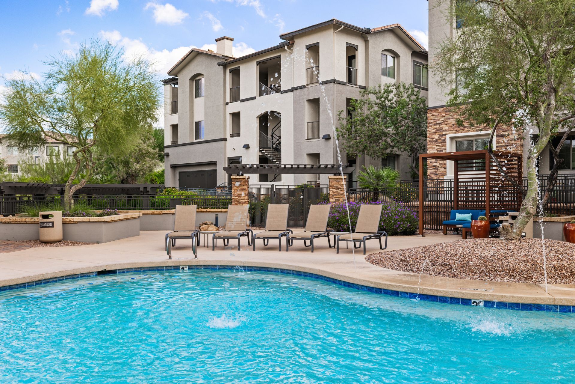 Apartment pool with lounge chairs and landscaped courtyard under a partly cloudy sky