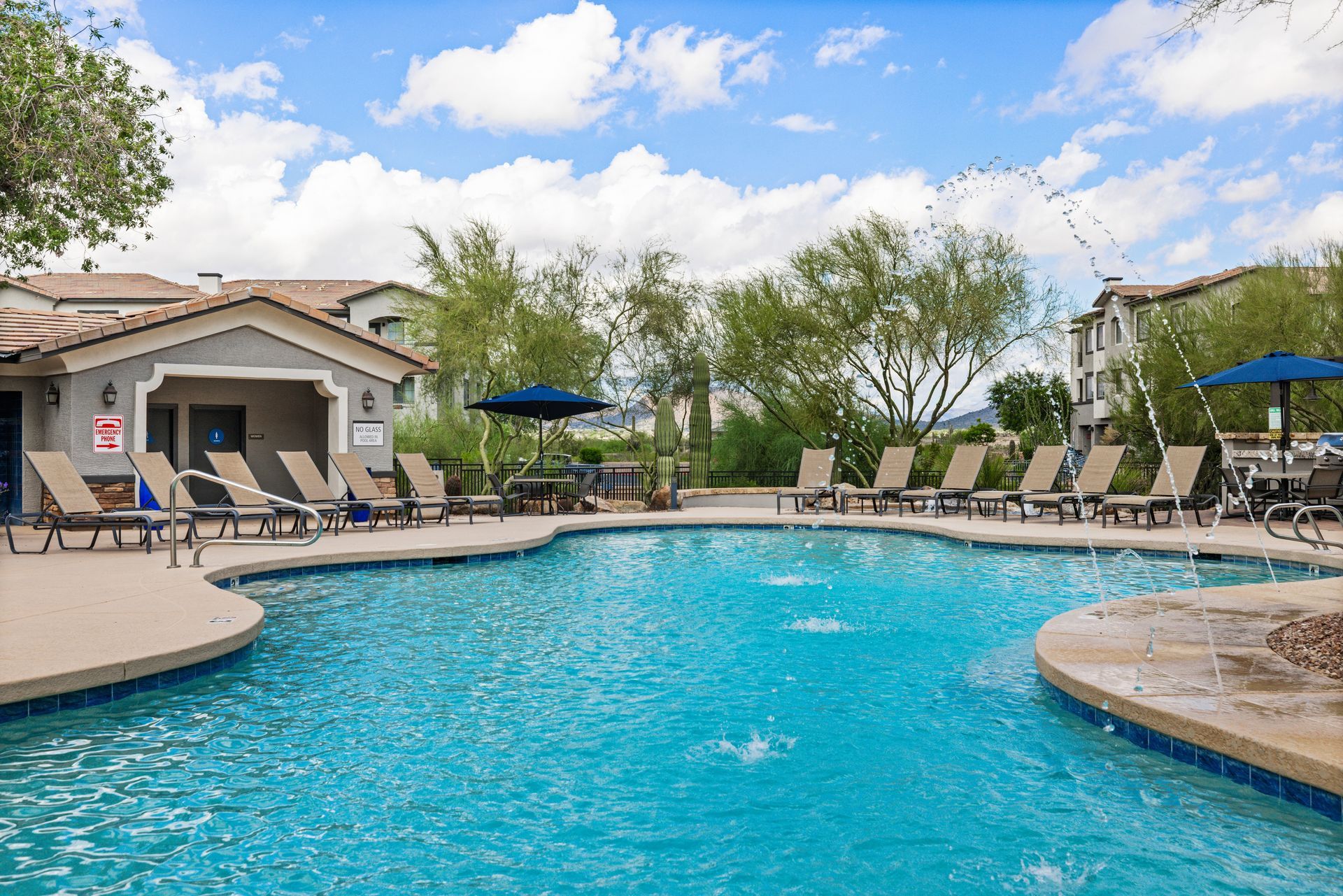 Outdoor apartment pool with blue water, lounge chairs, umbrellas, and a clubhouse under a partly cloudy sky
