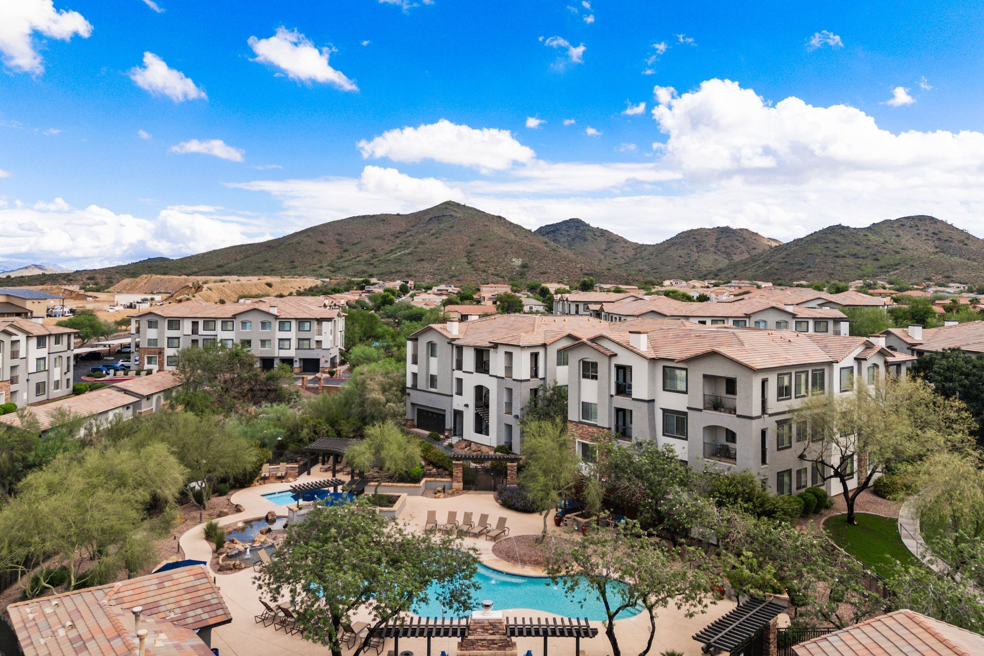 Aerial view of a suburban apartment complex with beige buildings under a bright blue, cloudy sky, and a pool