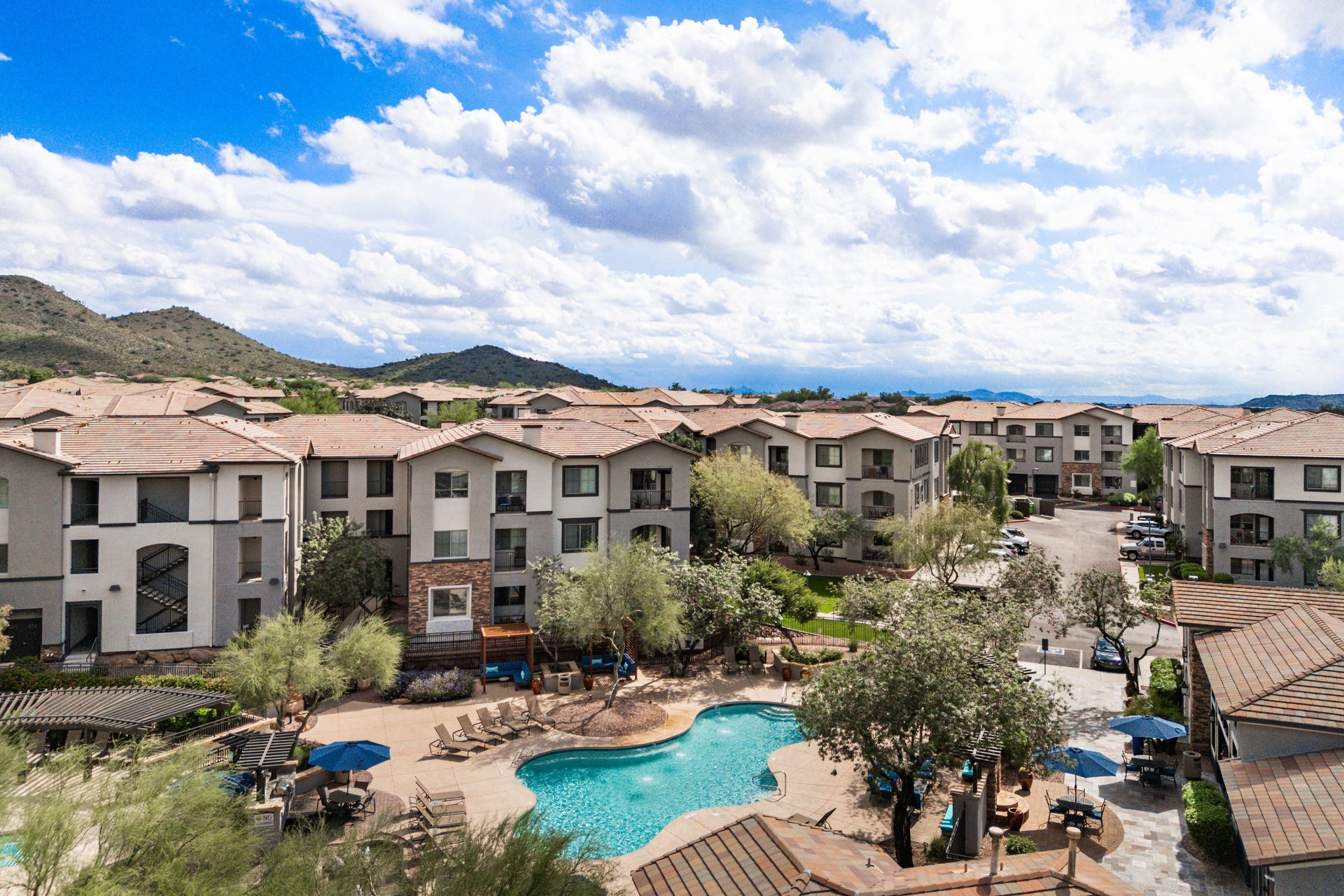 Apartment complex with a blue pool, palm trees, and mountains under a partly cloudy sky