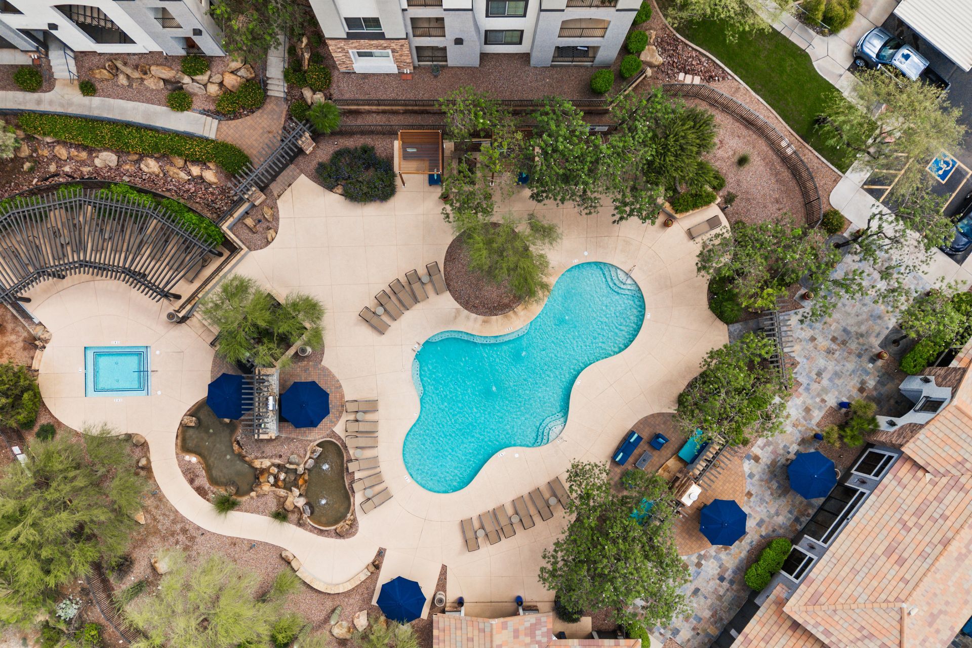 Aerial view of a resort pool area with a turquoise pool, lounge chairs, trees, and surrounding buildings