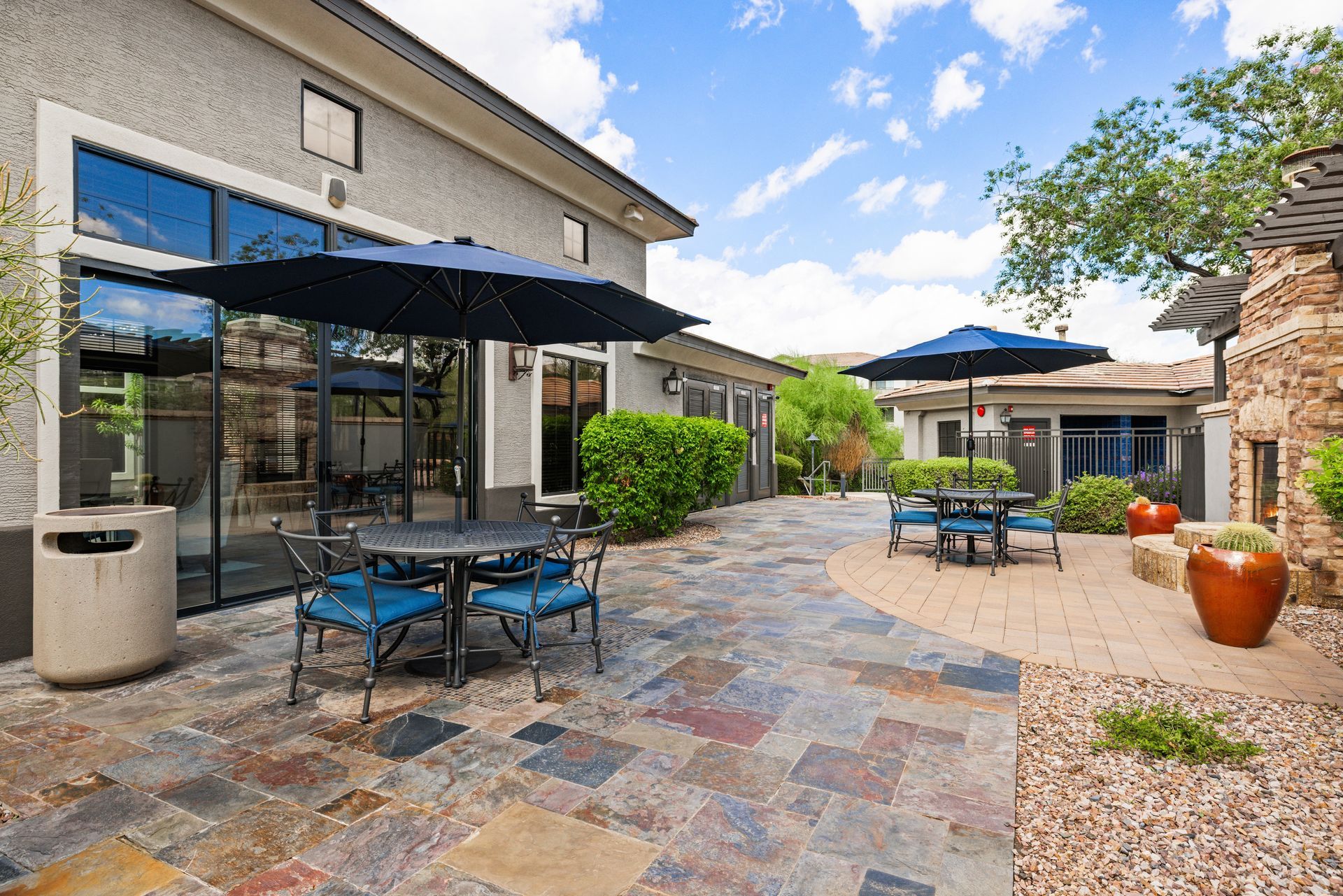Patio with stone paving, black umbrellas, and blue metal tables beside a building and landscaped courtyard