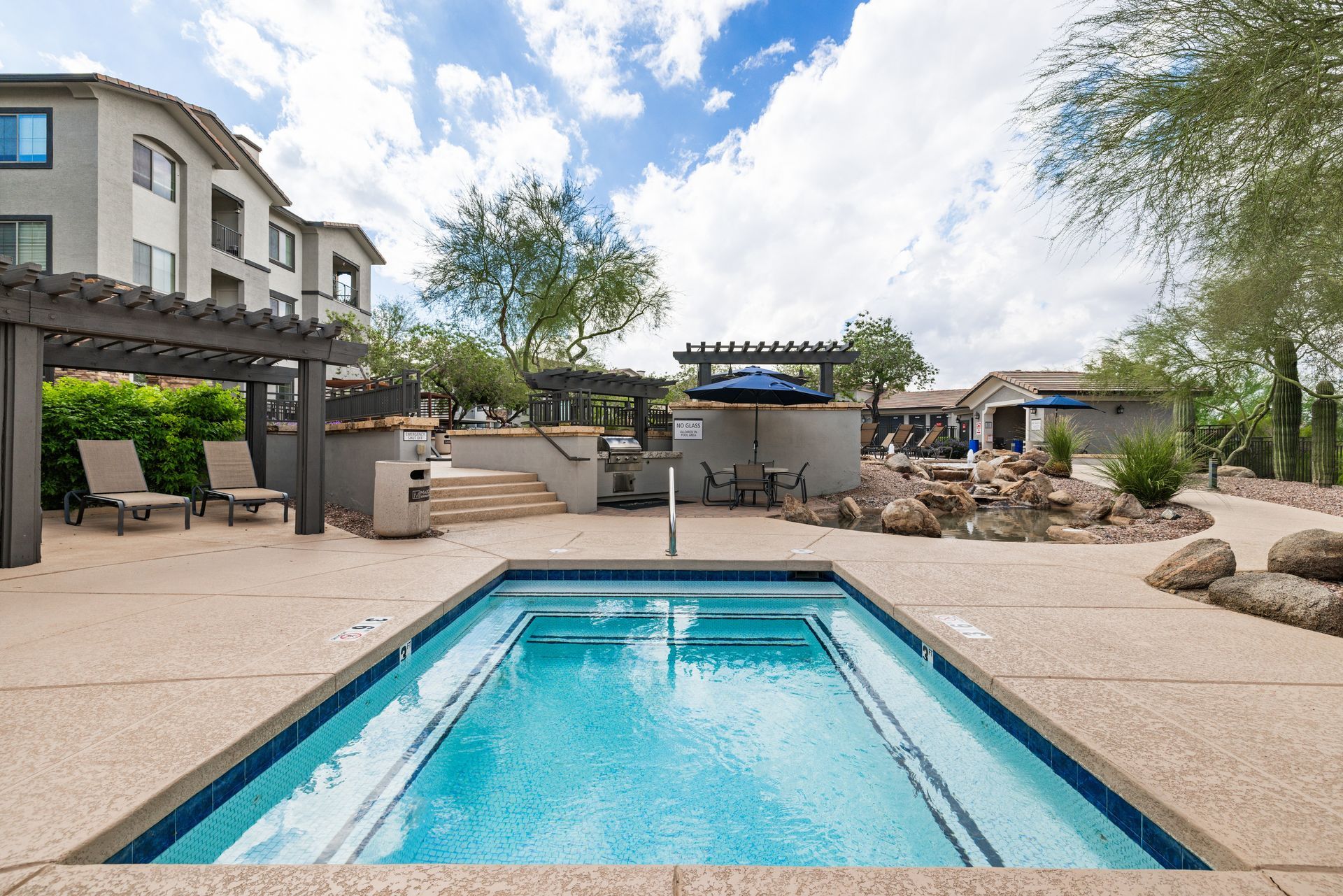 Backyard pool with lounge chairs, pergola seating, rocks, and a modern house under a partly cloudy sky