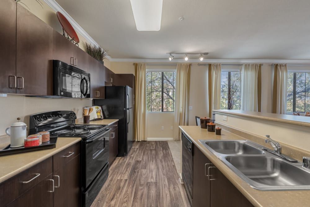 Modern kitchen with brown cabinets, black appliances, wood floors, and a double sink by a pass-through counter