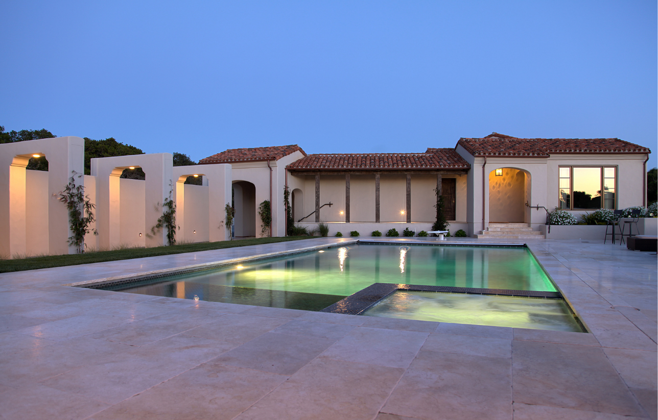 A luxurious, cream-colored home with a pool at dusk; terracotta roof, arches, and illuminated water.