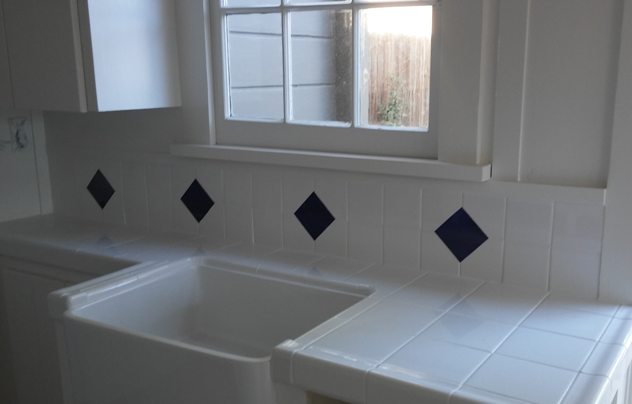 White kitchen with white sink and counter, tiled backsplash with blue diamond accents, window above sink.