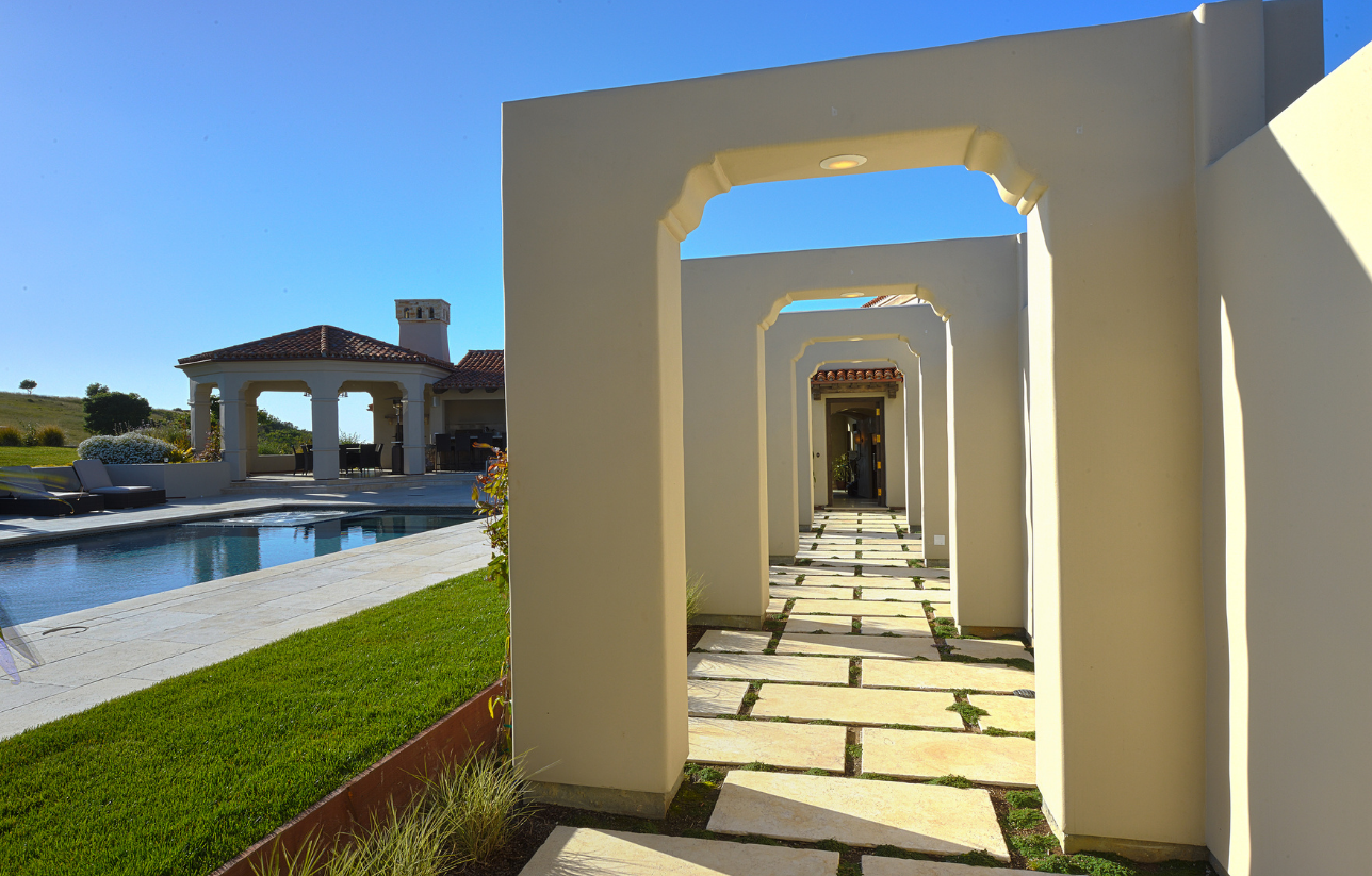 Stone path through archways leads to a pool and house on a sunny day.
