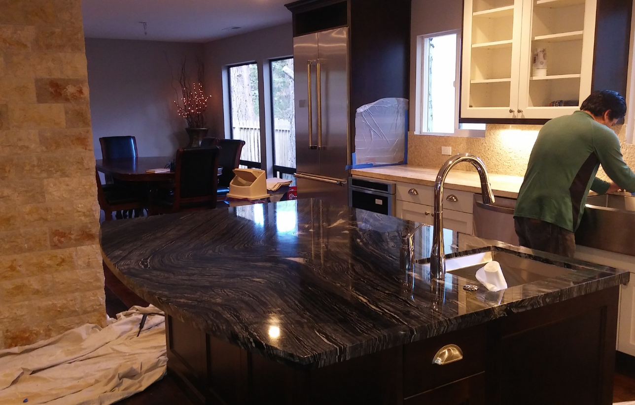 Kitchen island with a dark granite countertop, person by the sink.