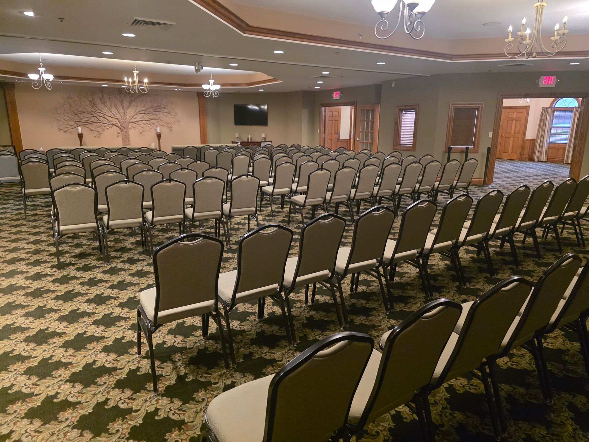 Empty banquet hall with rows of chairs facing a front area, patterned carpet, and chandeliers.