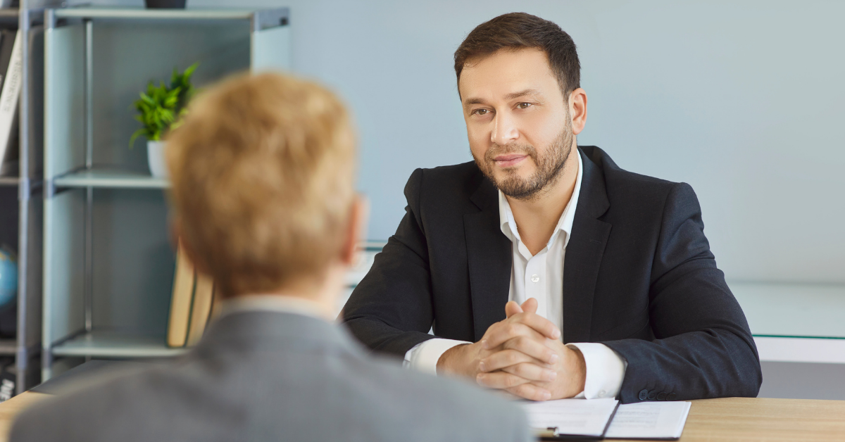 Two people in a business meeting across a desk, with one man speaking and smiling in an office.