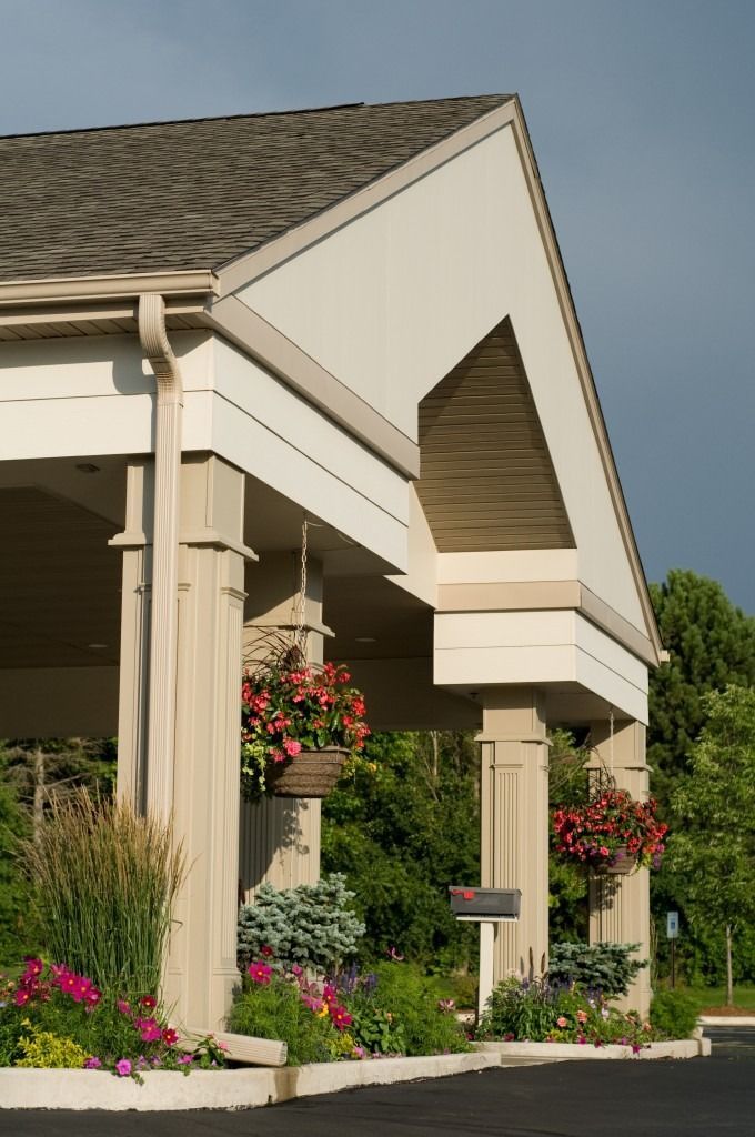 Exterior of a building entrance with beige columns, hanging flower baskets, and landscaped greenery.