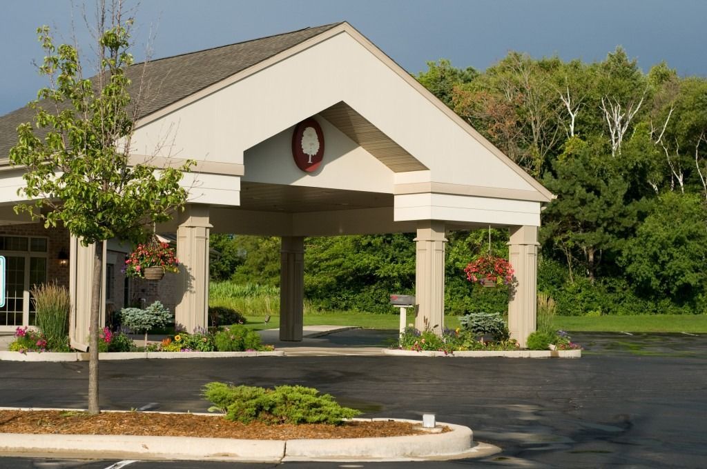 Covered building entrance with columns and a wreath, beside a paved driveway and trees.