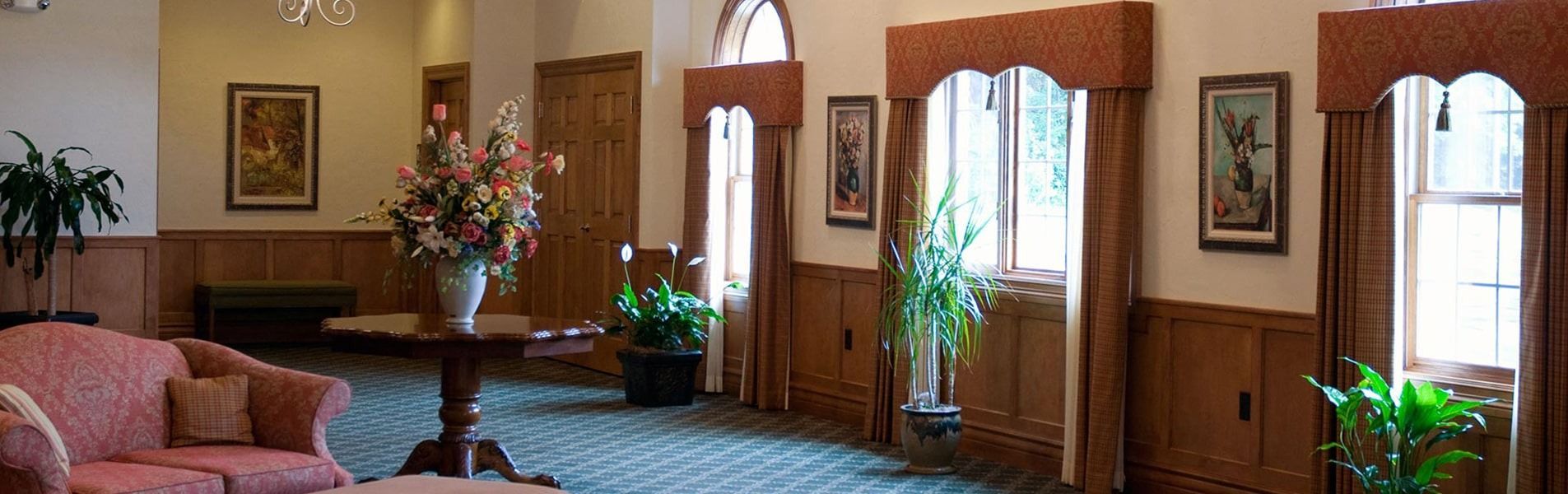 Elegant living room with pink sofa, patterned carpet, wooden trim, and tall windows with plants and curtains