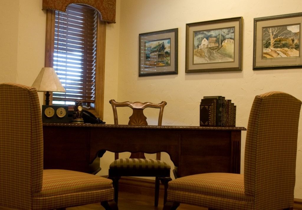 Warm office with two wicker chairs, wooden desk, table lamp, and framed landscape paintings on the wall.