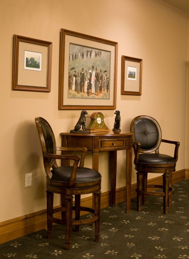 Two wooden chairs flank a small round table in a carpeted hallway with framed artwork on beige walls.
